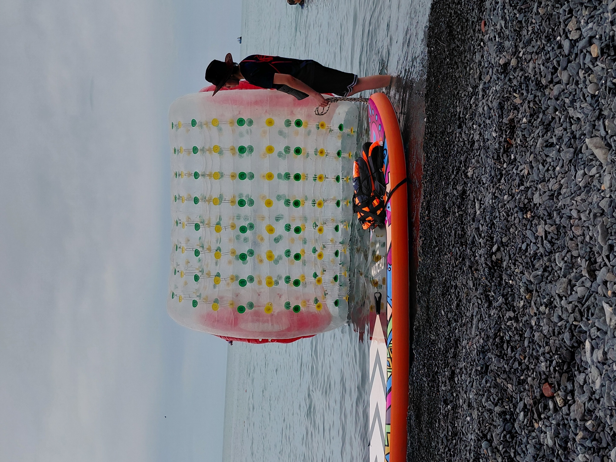 A person wearing a hat stands in the water next to an inflatable water roller. In the foreground, an orange and multi-colored paddleboard lies partially submerged on a rocky shore. The person is holding a rope attached to the water roller. The sky is overcast. This image does not show the city of Ақши, Kazakhstan.