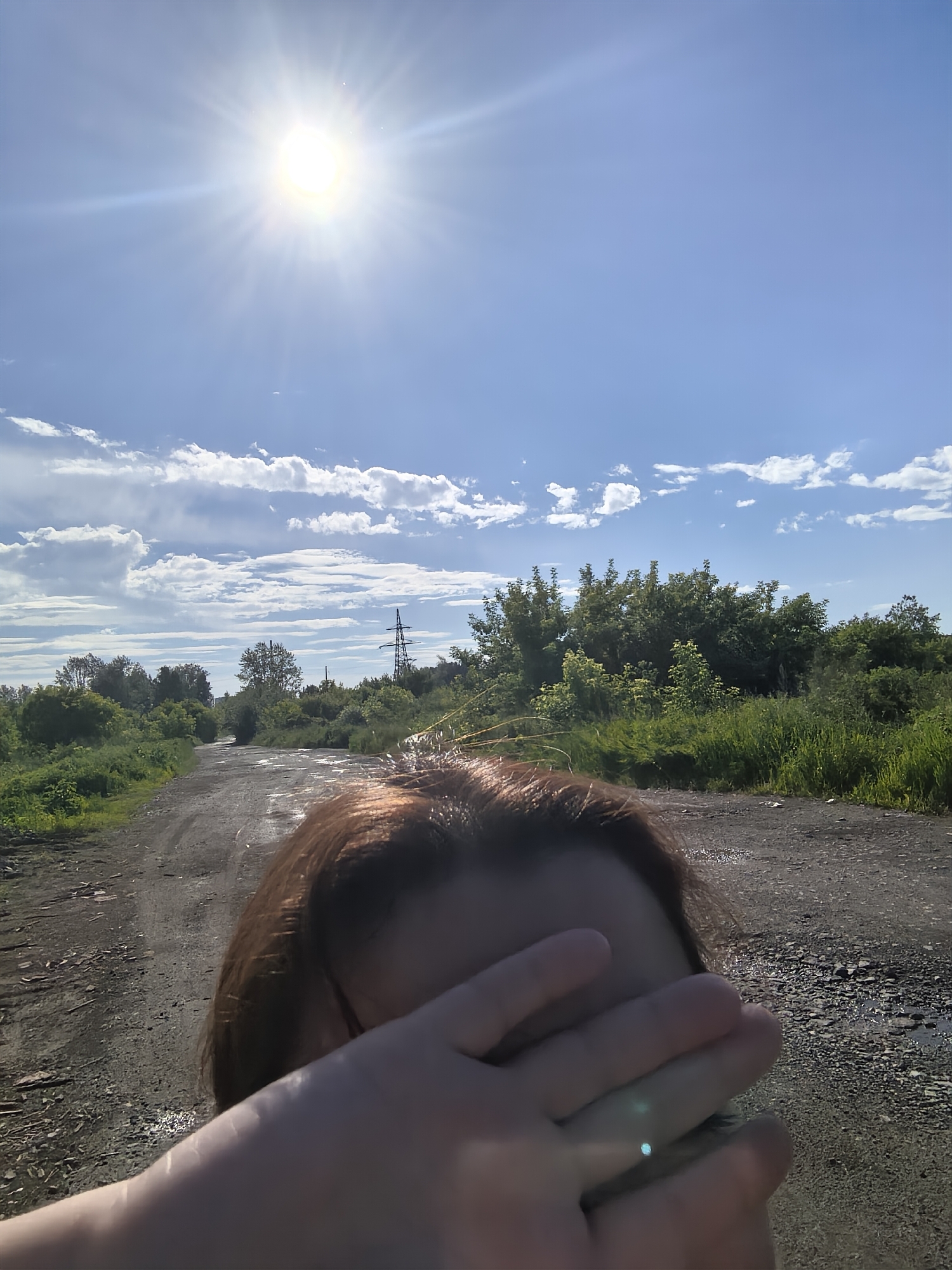 The image shows a person's head and hand covering their face, in the foreground. The person has brown hair and is obscuring their eyes with their hand. Behind them, a dirt road stretches into the distance, lined with green trees and vegetation. The sky is a bright blue with scattered white clouds, and the sun is shining intensely, creating a lens flare. A power line tower is visible in the middle ground on the right side of the image. The scene appears to be outdoors in a rural or undeveloped area, likely during the daytime due to the bright sunlight and clear sky. The presence of the sun flare suggests it is a sunny day.