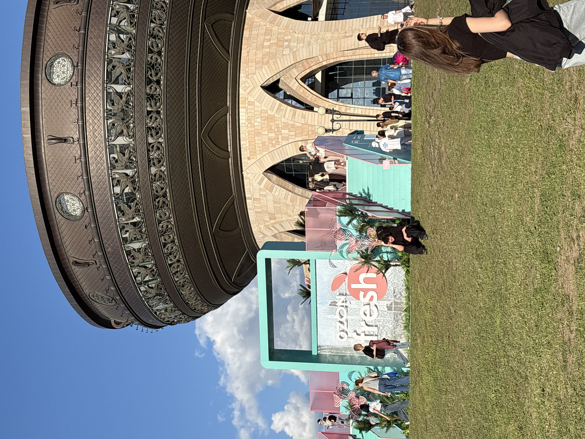This image captures the Kazan Family Center, also known as the Kazan Wedding Palace, in Kazan, Russia, under a clear blue sky with scattered white clouds. The prominent structure features a large, dark, bronze-toned, cauldron-like upper section, intricately detailed with a diamond-patterned texture, circular decorative medallions, and a band of relief sculptures depicting winged griffins or dragons. Below this, the building's base is constructed from light brown masonry, characterized by prominent pointed arches that form windows and entrances.
In the midground, on a vibrant green grassy slope, a temporary promotional installation for "Ozon fresh" is set up. This installation consists of multi-level teal-colored frames and platforms, accented with pink elements and stylized pink palm leaf cutouts. A central teal frame features a cascading waterfall effect, over which the "Ozon fresh" logo with an apple graphic is prominently displayed. Teal stairs lead up to a pink-railed platform, which people are ascending and descending.
Numerous individuals are dispersed throughout the scene, most casually dressed. In the immediate foreground on the right, a young woman with long brown hair, wearing a black t-shirt and light-colored jeans, with a black jacket draped over her arm, is partially visible from behind, observing the scene. To the left, a person dressed entirely in black sits on the grass.
A group of people is congregated around the "Ozon fresh" installation, with some walking along the base of the structure, others on the stairs, and a few on the upper platforms. More individuals are visible near the main entrance of the Kazan Family Center, walking up and down the building's steps and gathering at its base. The overall atmosphere appears sunny and active, suggesting a public event or a popular tourist destination.
