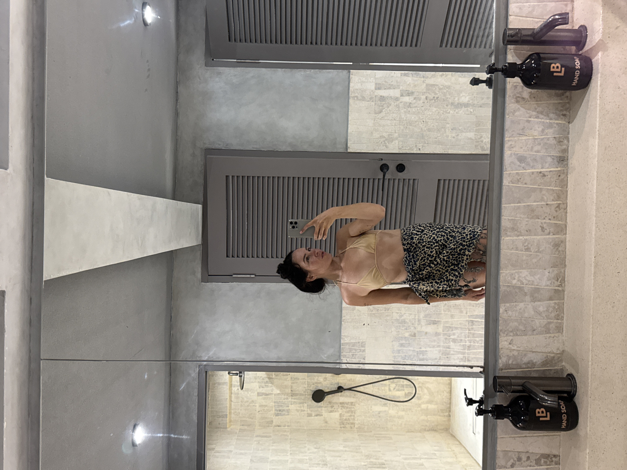 A woman stands in front of a large mirror in what appears to be a bathroom or changing area. She is holding a smartphone in her right hand, taking a selfie. She is wearing a light-colored bikini top and a leopard-print sarong tied around her waist. Her hair is pulled up into a bun. To her left, a shower stall with textured tiles is visible. To her right, a louvered door is present. The mirror reflects the entire scene, including the woman, the shower, and the door. Below the mirror, a countertop features two black soap dispensers and faucets. The overall environment appears to be modern with a focus on neutral tones and textures. This scene is located in North Kuta, Indonesia.