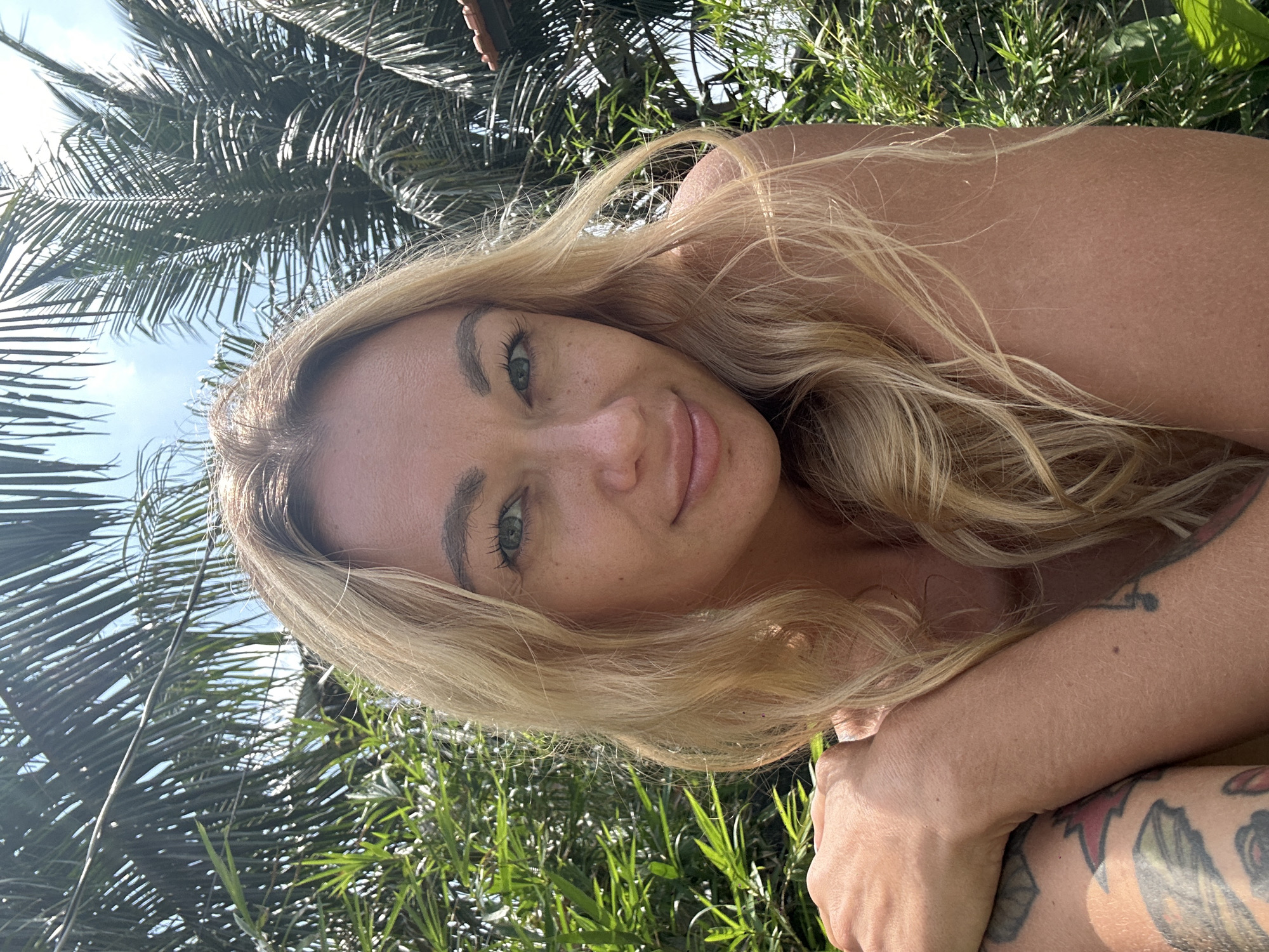 A blonde woman with blue eyes and fair skin is posing for a selfie outdoors. She is looking directly at the camera and smiling slightly. Her hair is long and wavy, and she is wearing a light-colored top that exposes her shoulders. She has tattoos on her arms, one of which is partially visible on her left arm. The background of the image consists of lush green foliage, including palm trees, suggesting a tropical location. The lighting indicates it is daytime, with bright sunlight casting shadows. The woman's pose, with her arm resting on her chest and her head tilted slightly, conveys a relaxed and casual demeanor. The scene is consistent with being in a garden or resort area in Mũi Né, Vietnam, known for its beaches and palm tree-lined landscapes.