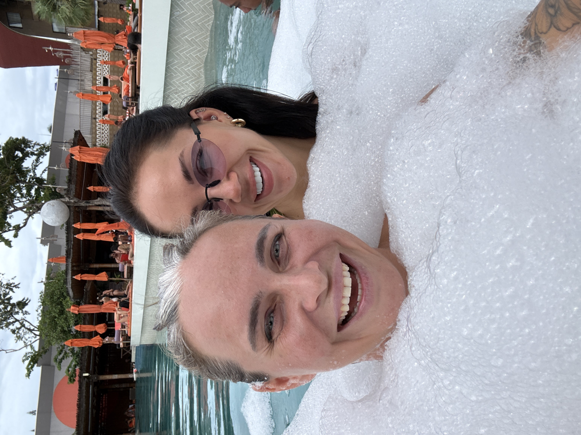 Two women are in a bubble-filled hot tub at a resort in Koh Samui District, Thailand. In the foreground, a woman with grey-streaked hair and blue eyes smiles broadly at the camera. Her mouth is open, revealing white teeth. To her right, partially obscured by bubbles and her companion, is another woman with dark hair, wearing heart-shaped pink sunglasses and gold hoop earrings. She also appears to be smiling. Behind them, the pool area of the resort is visible, with lounge chairs and orange umbrellas. The sky is overcast.
