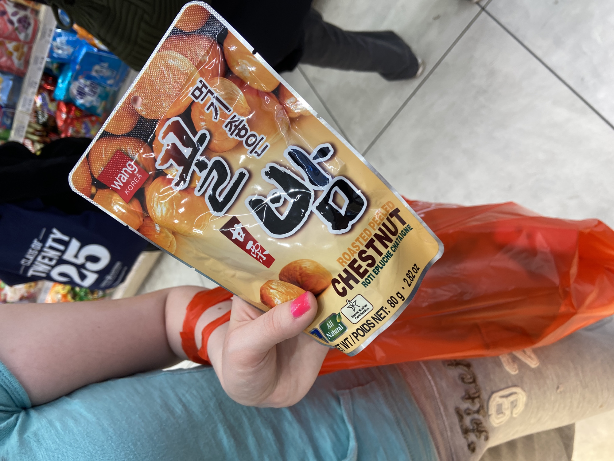A person is holding a package of "Wang Korea" roasted peeled chestnuts. The package is predominantly beige and orange, with Korean characters in large black font. The text "Roasted Peeled Chestnut" is displayed in English and French. The person has light skin, pink nail polish, and is wearing a light blue top, grey sweatpants, and an orange bracelet or wristband. The background shows a store aisle with shelves of items, a blue t-shirt with "Class of Twenty 25", and an orange shopping bag. The location appears to be inside a store.