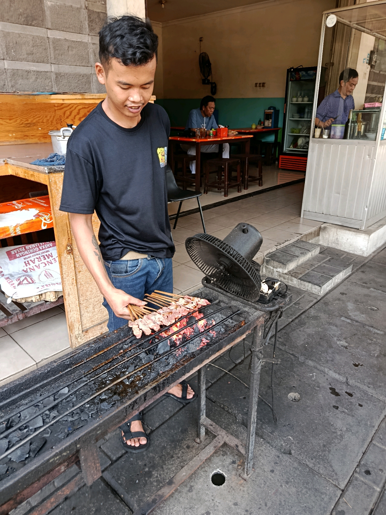 In Pusakarakyat, Indonesia, a young man wearing a black t-shirt and blue jeans stands in front of a charcoal grill, arranging skewers of meat. The grill is hot, with glowing embers visible. A black fan is placed on the side of the grill. In the background, a man sits at a table in an eatery, and another man is visible behind a glass counter. The environment appears to be an outdoor dining area of a restaurant or food stall.