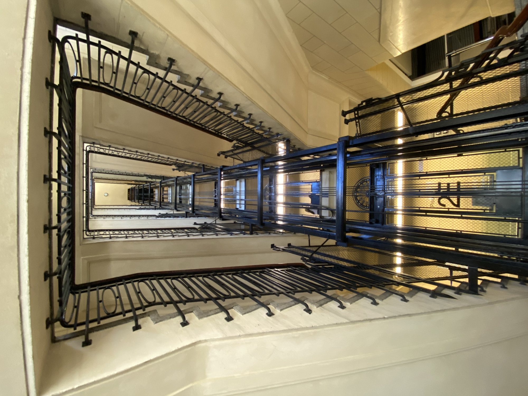 The image captures a view looking upward inside an elevator shaft in Buenos Aires, Argentina. The shaft is surrounded by a staircase with black wrought-iron railings, creating a visual frame for the elevator mechanics. The elevator cage's metal framework is visible, with mesh panels and signage indicating floor numbers. Light filters through the shaft, creating highlights and shadows on the metal and the off-white walls. The perspective is from the base of the shaft, emphasizing the verticality and depth of the space.