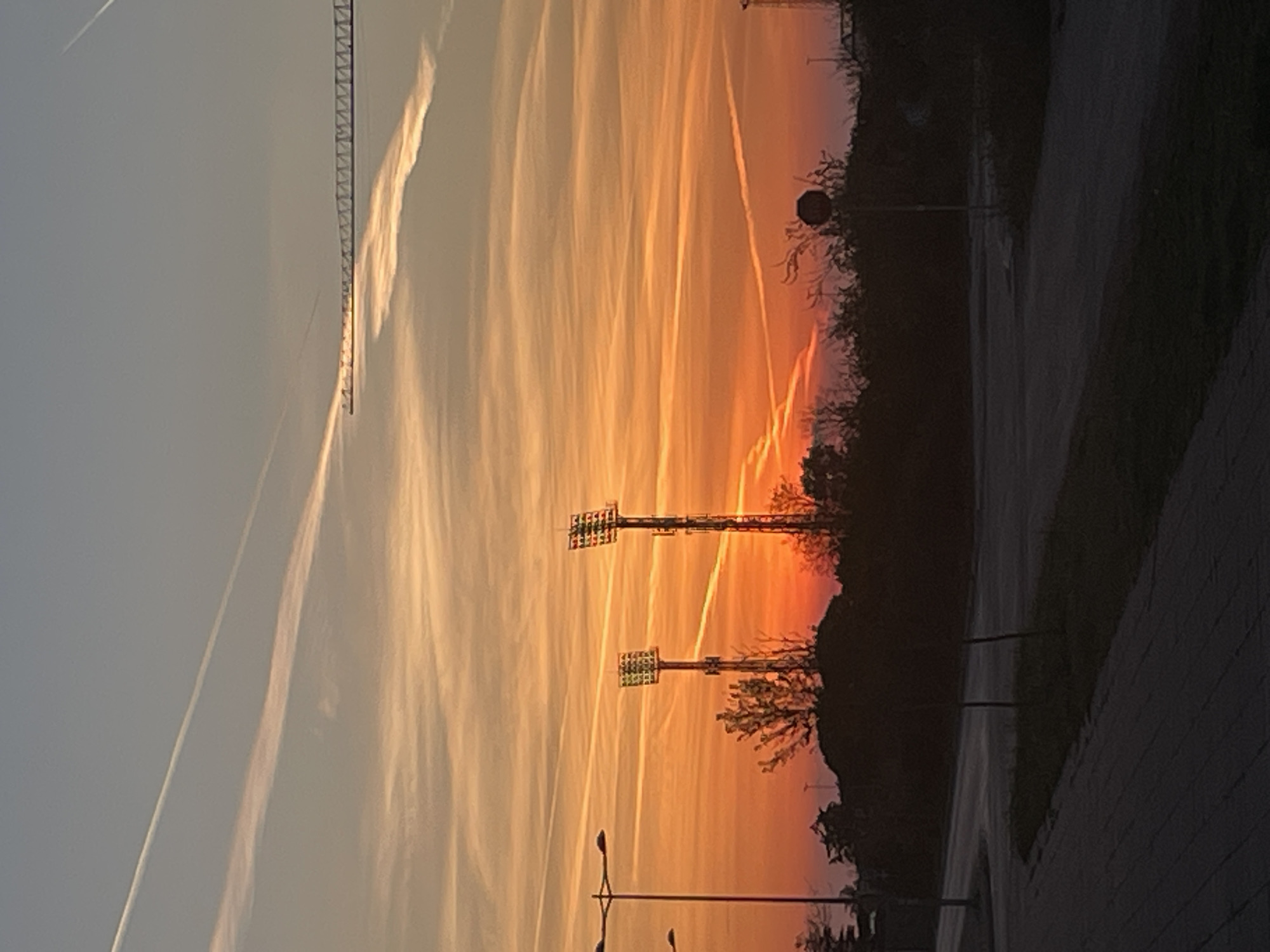 The image shows a sunset in Bar, Montenegro. The sky is orange and yellow with contrails from airplanes. There is a crane in the upper left of the image and two sets of floodlights. There are trees and other foliage silhouetted against the sunset. The right side of the image shows a sidewalk and street. A stop sign is also present.