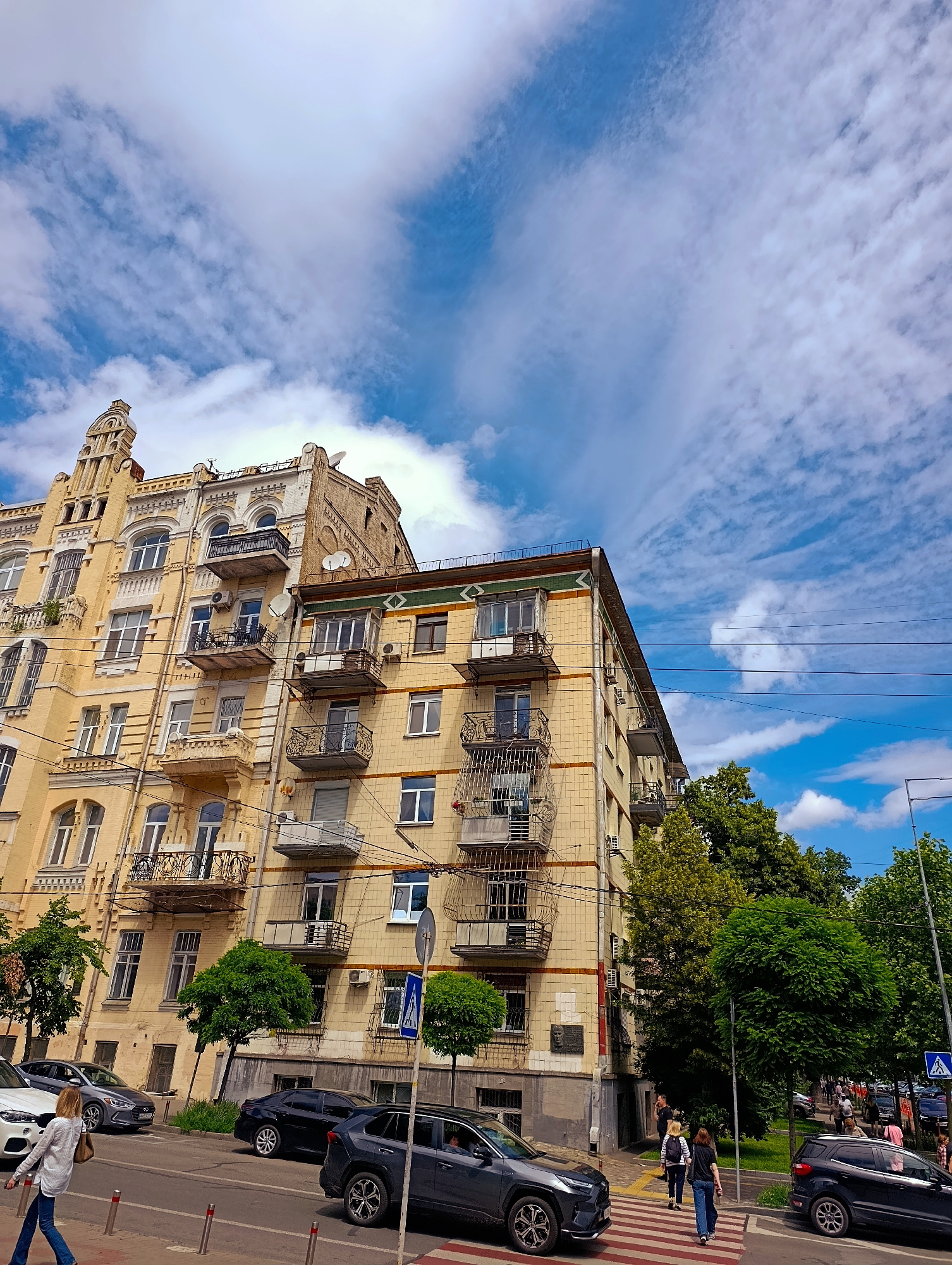 The image captures a street scene in Kyiv, Ukraine, dominated by a tall, yellow-hued building with intricate architectural details and numerous balconies. The building appears to be an apartment complex, with many windows and signs of habitation. In front of the building, a street runs parallel, with several cars parked and driving. A pedestrian crossing is visible in the foreground, with a woman walking across it. Lush green trees line the street and partially obscure the lower floors of the building. The sky above is a vibrant blue, dotted with fluffy white clouds. The overall impression is of a lively urban environment on a bright day.