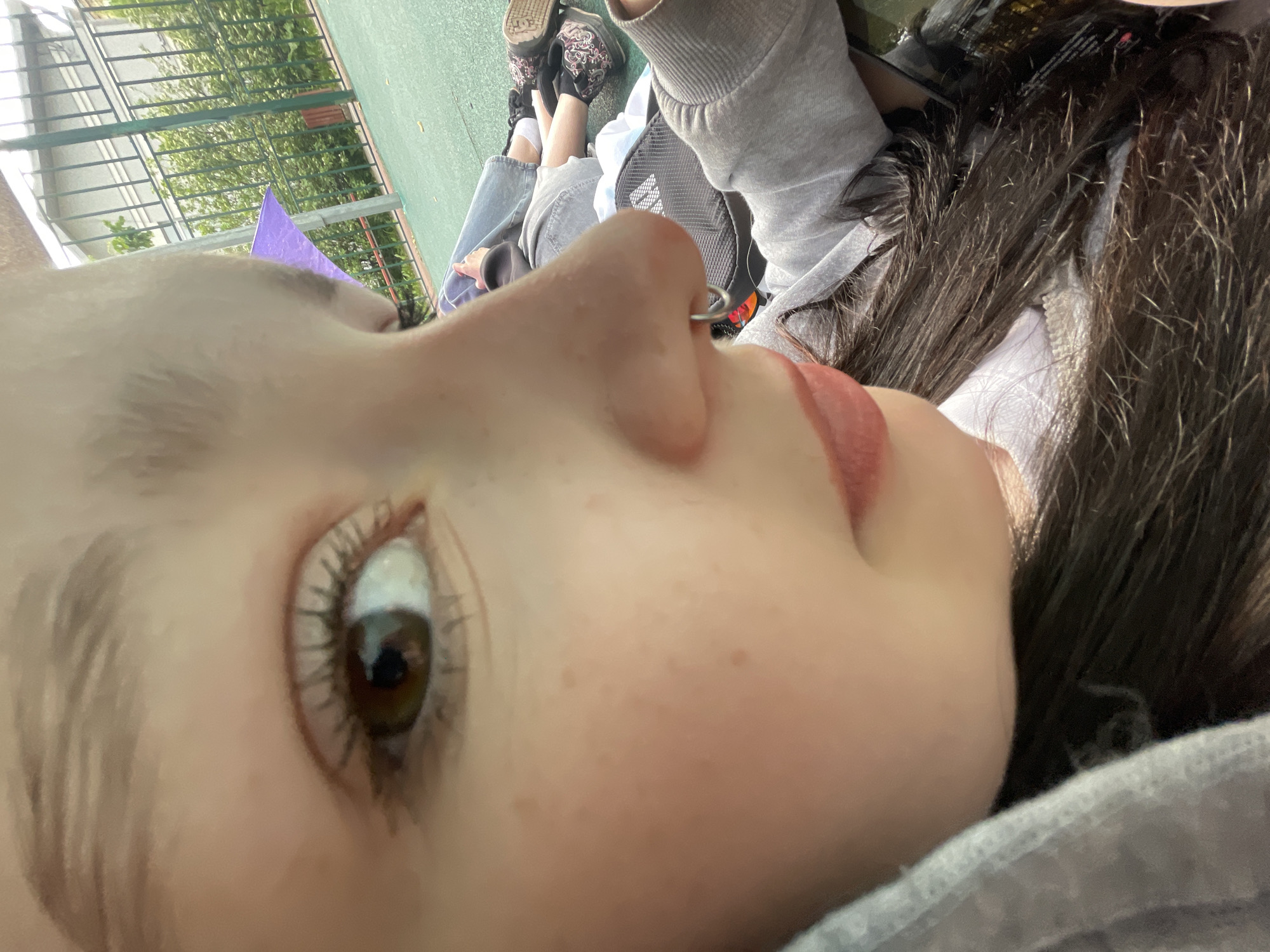 The image captures a close-up, side profile of a young person, rotated 90 degrees counter-clockwise, suggesting they are lying down or leaning back. The subject's face is prominent, showcasing light-toned skin, a brown eye with defined eyelashes, and a nose featuring a small silver hoop piercing. Their lips are painted with a light pink lipstick. Dark, long hair frames the side of their face. They are dressed in a grey hooded sweatshirt and light blue jeans, with black and white sneakers visible further in the background. A black backpack with a partially visible "ADIDAS" logo rests near them.
The setting appears to be outdoors during the daytime. The person is situated on a green ground surface, resembling a sports court or artificial turf. In the background, there is a metal fence or structure, and beyond it, green trees and foliage are visible. A purple object, possibly a piece of fabric or paper, is partially seen near the top of the subject's head. The lighting is bright and even, consistent with daylight. The scene conveys a casual and relaxed atmosphere. The provided location context is Saint Petersburg, Russia.