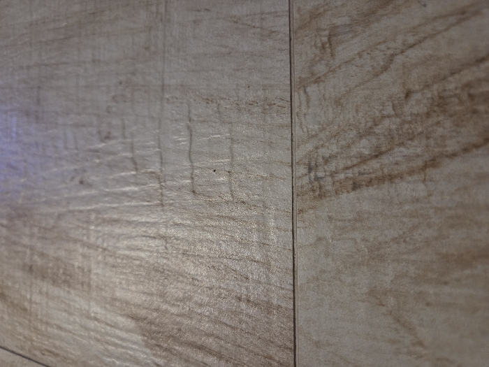 The close-up shot features three adjacent wall tiles. The tiles exhibit a wood-grain pattern in shades of beige and brown. The leftmost tile has a lighter tone with streaks of darker brown, while the tile on the right is dominated by more pronounced brown patterns. A vertical line separates the tiles indicating grout or a joint, emphasizing the uniformity and texture of the tiled surface.
