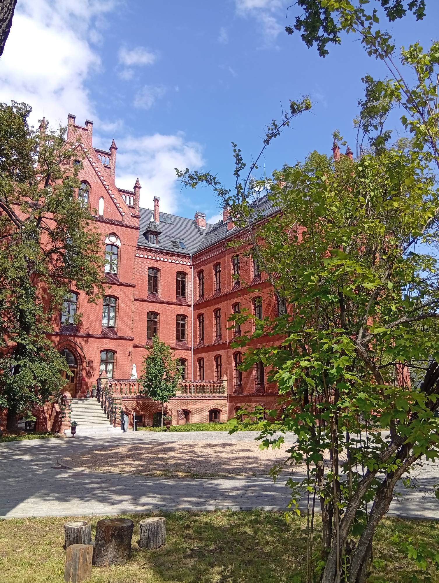 The image depicts a red brick building with multiple stories and a gabled roof, characterized by a classical architectural style. The building features numerous arched windows with dark frames and decorative brickwork accents along the facade. A wide, paved courtyard is situated in front of the building, with a section of gravel in the center. A set of stairs leads up to the main entrance, flanked by railings and planters.
To the left of the building, a large tree with dense green foliage partially obscures the structure. In the foreground, several tree stumps are arranged on a grassy area. A young tree with lighter green leaves stands in the mid-ground on the right side of the image, also partially obscuring the building. The sky above is blue with scattered white clouds.
This building is located at the Wrocław University of Science and Technology, specifically the **building of the Faculty of Civil Engineering at 25 Wybrzeże Wyspiańskiego in Wrocław, Poland**.