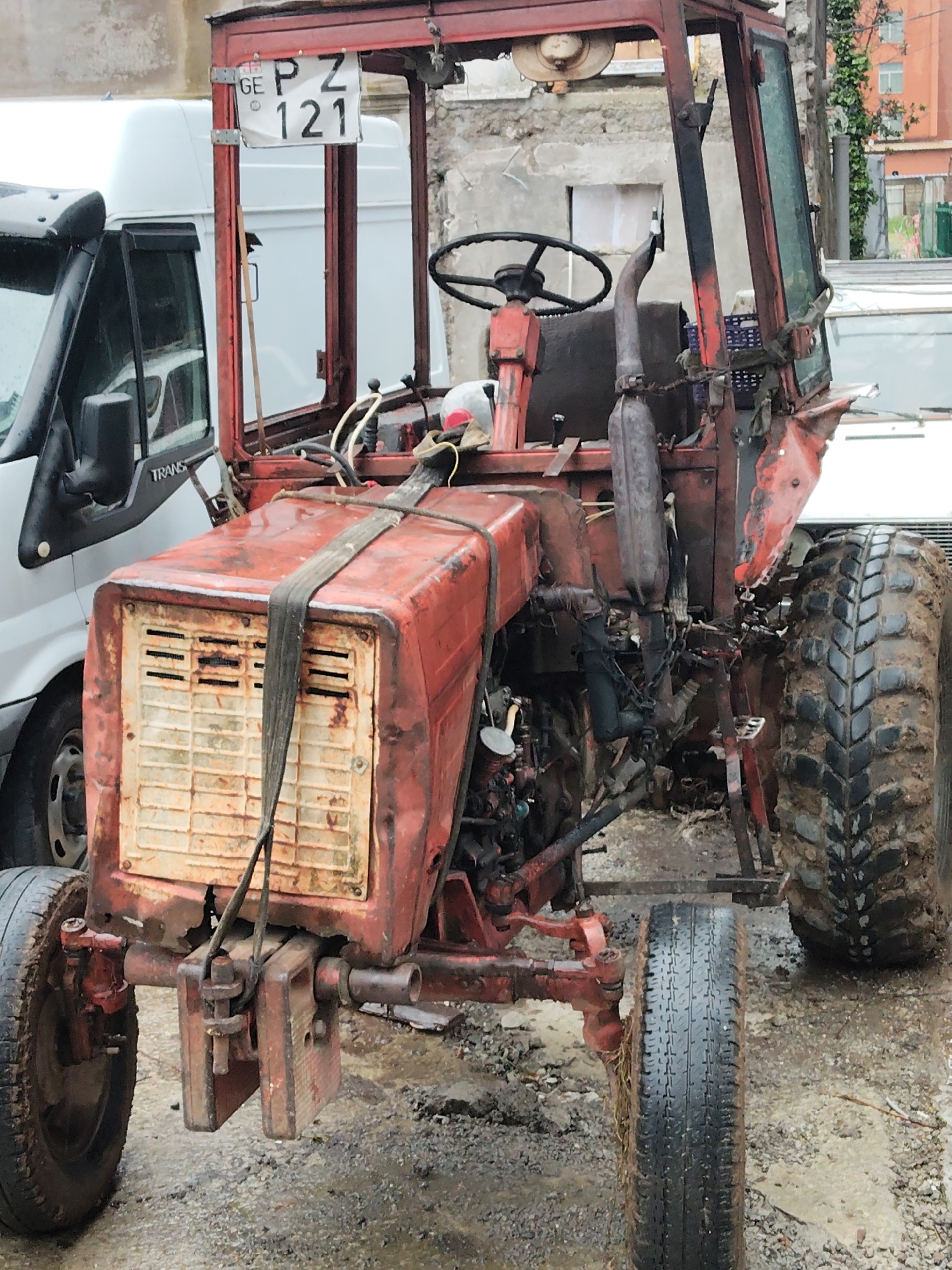 A weathered, red tractor with the license plate "GE PZ 121" is parked on a wet, possibly paved surface. The tractor is positioned in front of a white Ford Transit van. The environment appears to be urban, possibly in the city of Batumi, Georgia, judging by the "GE" on the license plate. The tractor appears to be old and is possibly in a state of disrepair. The front of the tractor has a strap securing the hood. The background includes a building with exposed concrete and windows with greenery.