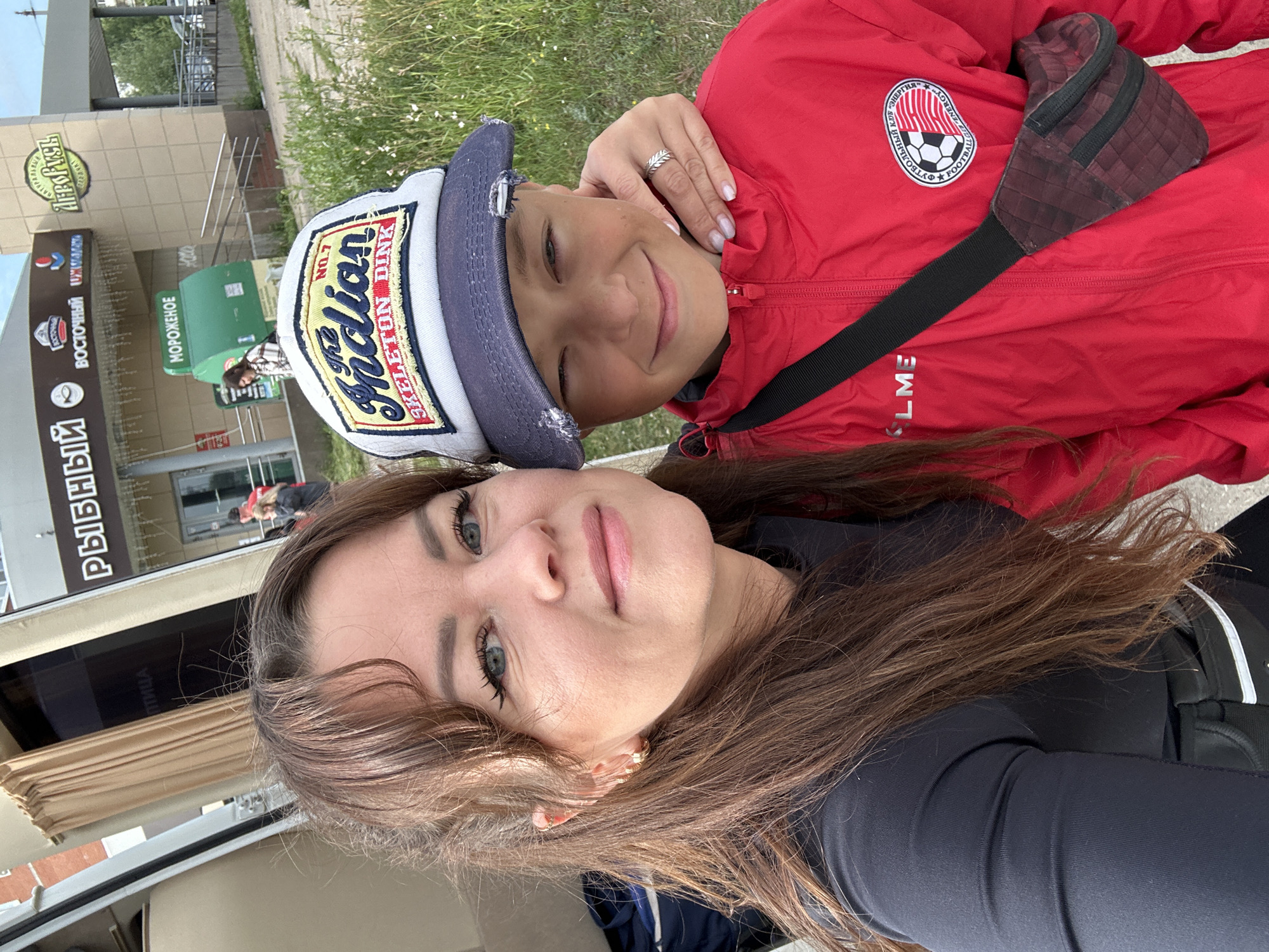 This is a close-up, selfie-style outdoor photograph taken in the city of Chaikovsky, Russia. A woman with brown hair and light-colored eyes is in the foreground, smiling at the camera. She is wearing a black long-sleeved shirt and gold hoop earrings. A young boy, wearing a red jacket and a baseball cap, is positioned next to her. The boy's right arm is draped over his shoulder. The woman's hand rests on the boy's shoulder, and she is wearing a ring on her right hand. The background shows a building with signage including "РЫБНЫЙ" (Fish) and "МОРОЖЕНОЕ" (Ice Cream), suggesting commercial establishments. There is also greenery and a paved area, indicative of an urban setting. The image appears to have been taken during daylight hours.