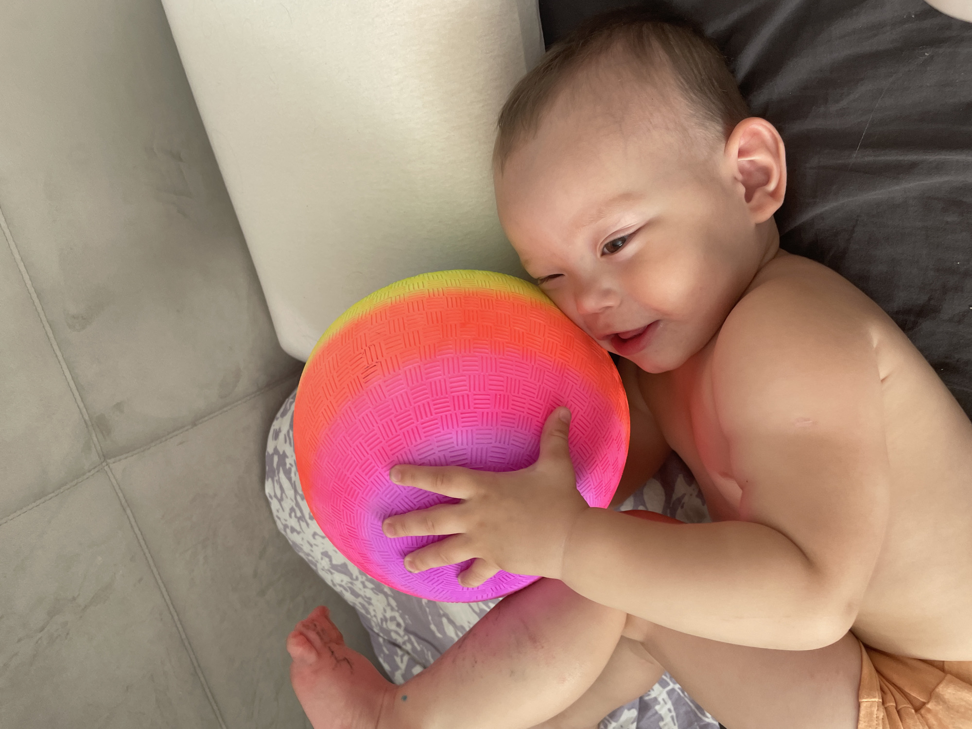 A baby is lying on their back on a bed, hugging a colorful, striped ball. The baby is light-skinned with brown hair and is wearing an orange diaper. The bed has a gray blanket and a white headboard. The floor is gray tile. The scene appears to be indoors. There is no information available in the image to confirm a specific location, such as Perm, Russia.