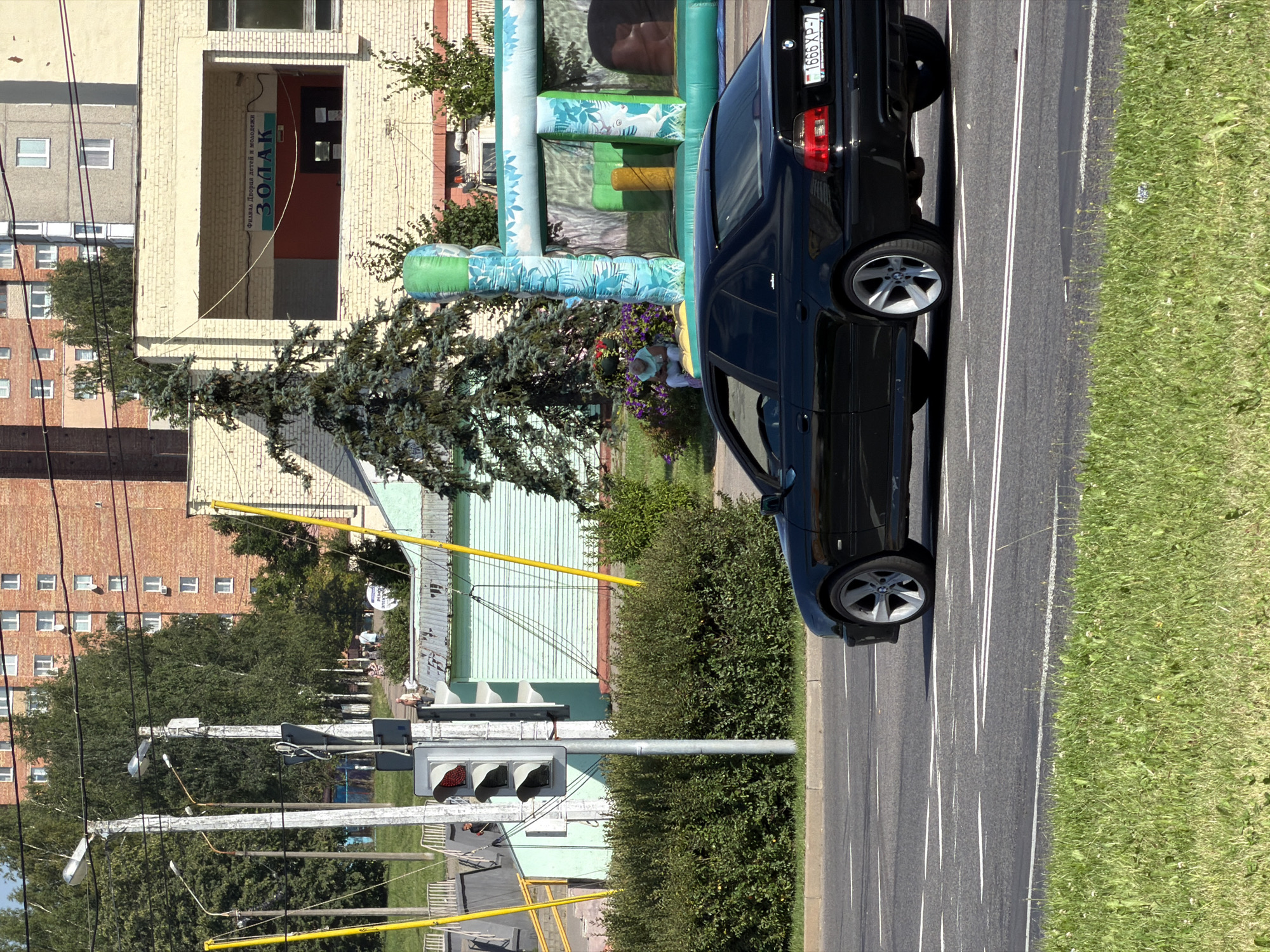 A black BMW sedan is driving on a street in Minsk, Belarus. The car is in the right lane of a two-lane road with white lane markings. The car is facing away from the camera. In the background, there are buildings and trees. There is also a traffic light and a pedestrian crossing. The weather is sunny.