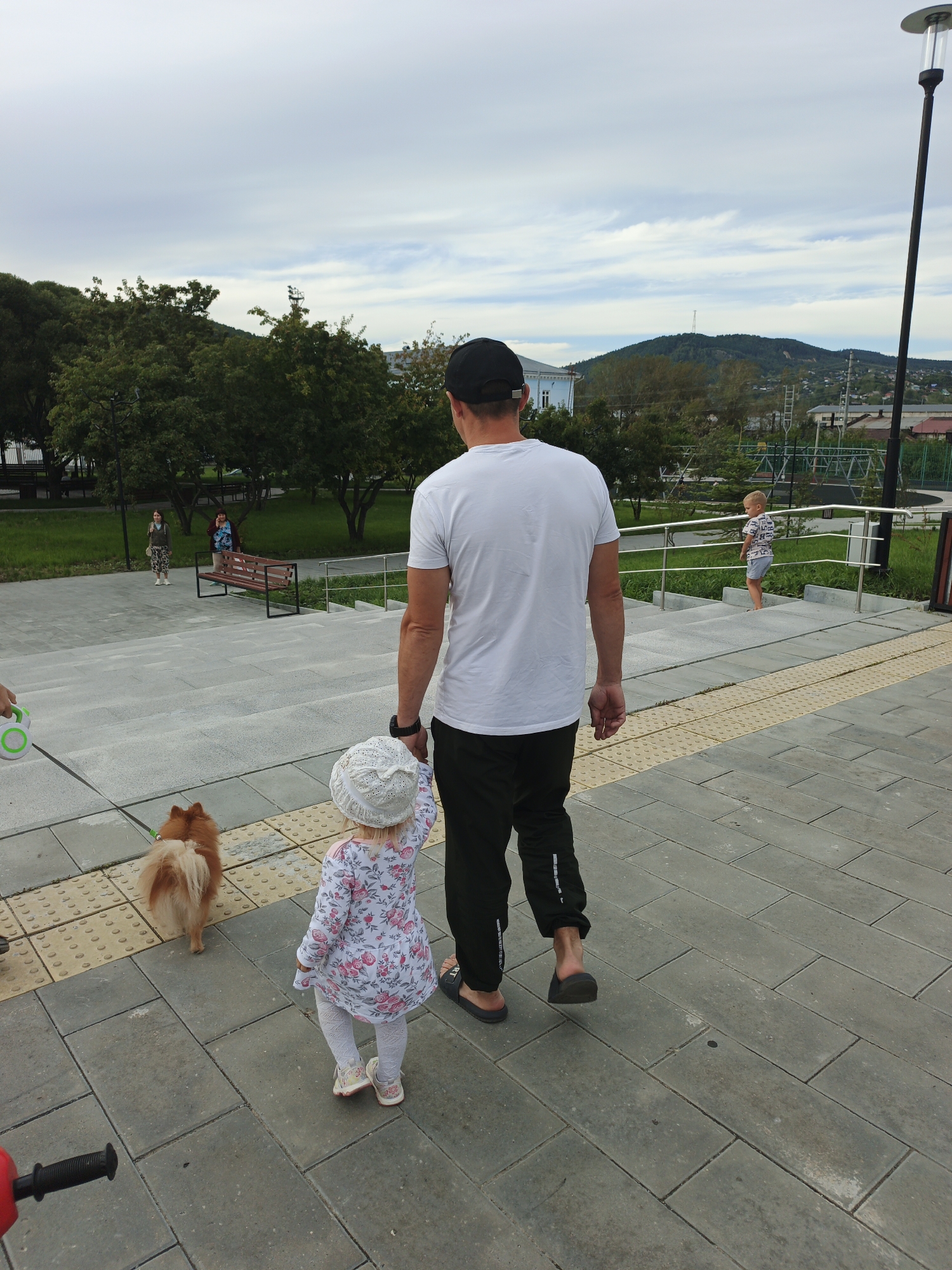In Zlatoust, Russia, on a paved outdoor area with grey square and rectangular paving stones, a man in a white t-shirt and black pants, wearing a black baseball cap and dark sandals, walks away from the camera. He is accompanied by a young child, who is facing away from the camera and wearing a white hat with a knitted texture, a white and pink floral dress, white tights, and light-colored shoes. To their left, a small, fluffy orange-brown dog on a leash, held by an unseen person, also walks away from the camera. In the background, there are stairs, benches, trees with green foliage, and hills with buildings and a communication tower. Other people are visible in the background, including a child on the stairs and two women standing near a bench.