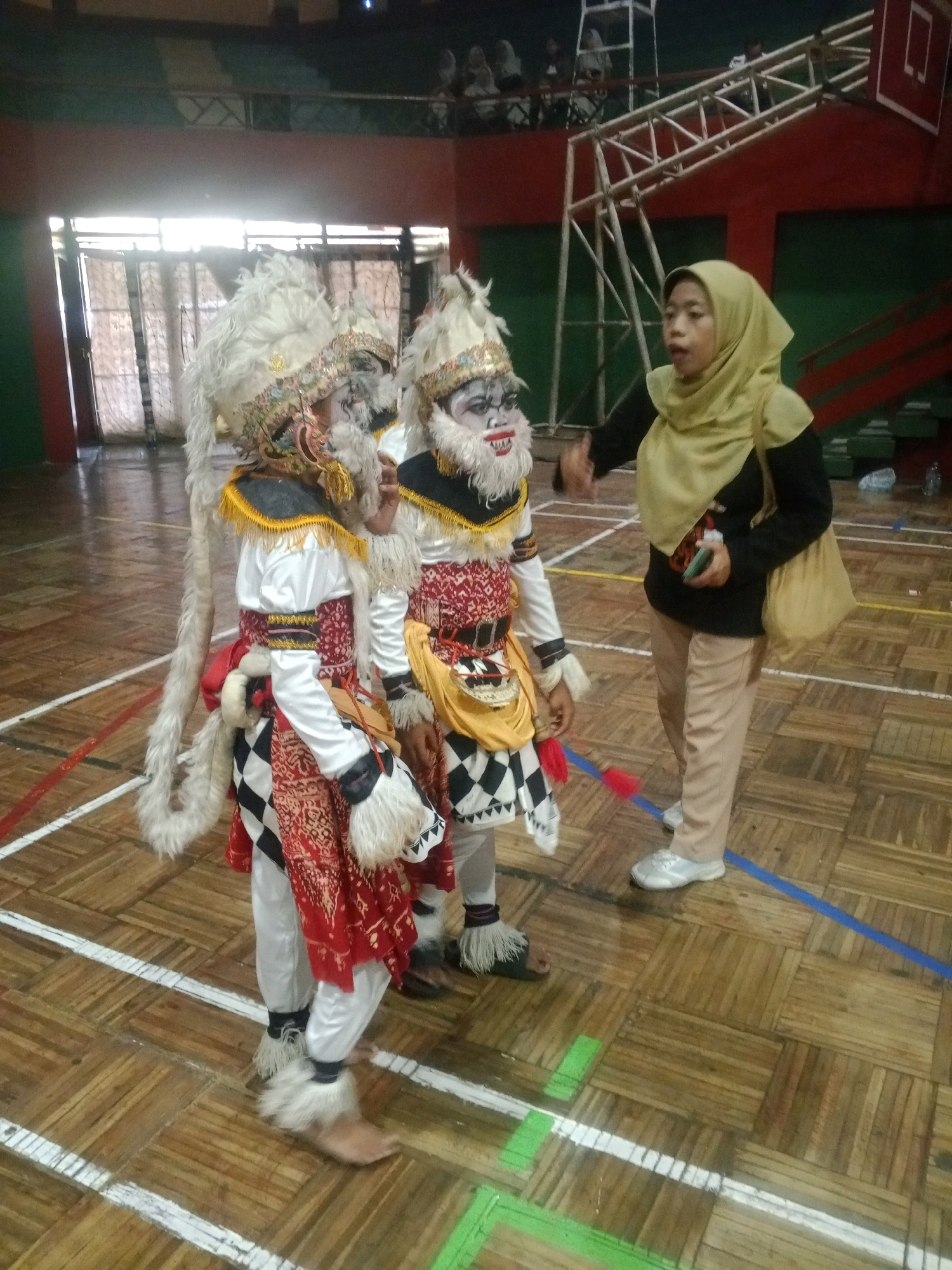 In Wuryorejo, Indonesia, within an indoor sports facility featuring a wooden parquet floor marked with white and blue lines, two individuals dressed in traditional performance costumes stand on the left. They wear elaborate headdresses with white fur, face paint resembling a white monkey mask with red accents, and outfits consisting of white shirts, red and black patterned vests, and white pants adorned with white fur trim around the ankles. One performer has a long, white, furry tail extending from the back of their costume. A woman in a light yellow hijab, black long-sleeved shirt, tan pants, and silver sneakers stands to the right, facing the performers. She holds a dark-colored smartphone in her right hand and has a light-colored shoulder bag on her right side. The background shows large windows with white curtains, metal scaffolding, and red walls, indicating an indoor public space. The lighting is bright, originating from the windows.
