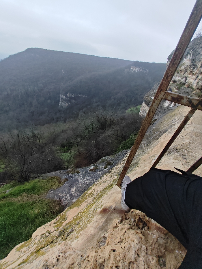 The image displays a first-person perspective from a rocky cliff edge within the Shumen Plateau region near Shumen, Bulgaria, under an overcast sky. In the immediate foreground on the lower right, a person's right leg, clad in dark trousers and a grey athletic shoe with a white sole, is positioned on a rough, light-brown rock surface mottled with green moss and lichen. Extending diagonally upwards from the lower right, a rusty metal ladder-like railing or structure is visible, anchored to the rock face. Below and to the left, a steep descent into a densely forested valley is depicted, featuring numerous bare deciduous trees and patches of green undergrowth. In the background, a large, tree-covered mountain or plateau spans the horizon, with visible rock outcrops on its slopes, all set against a uniformly grey, cloudy sky.