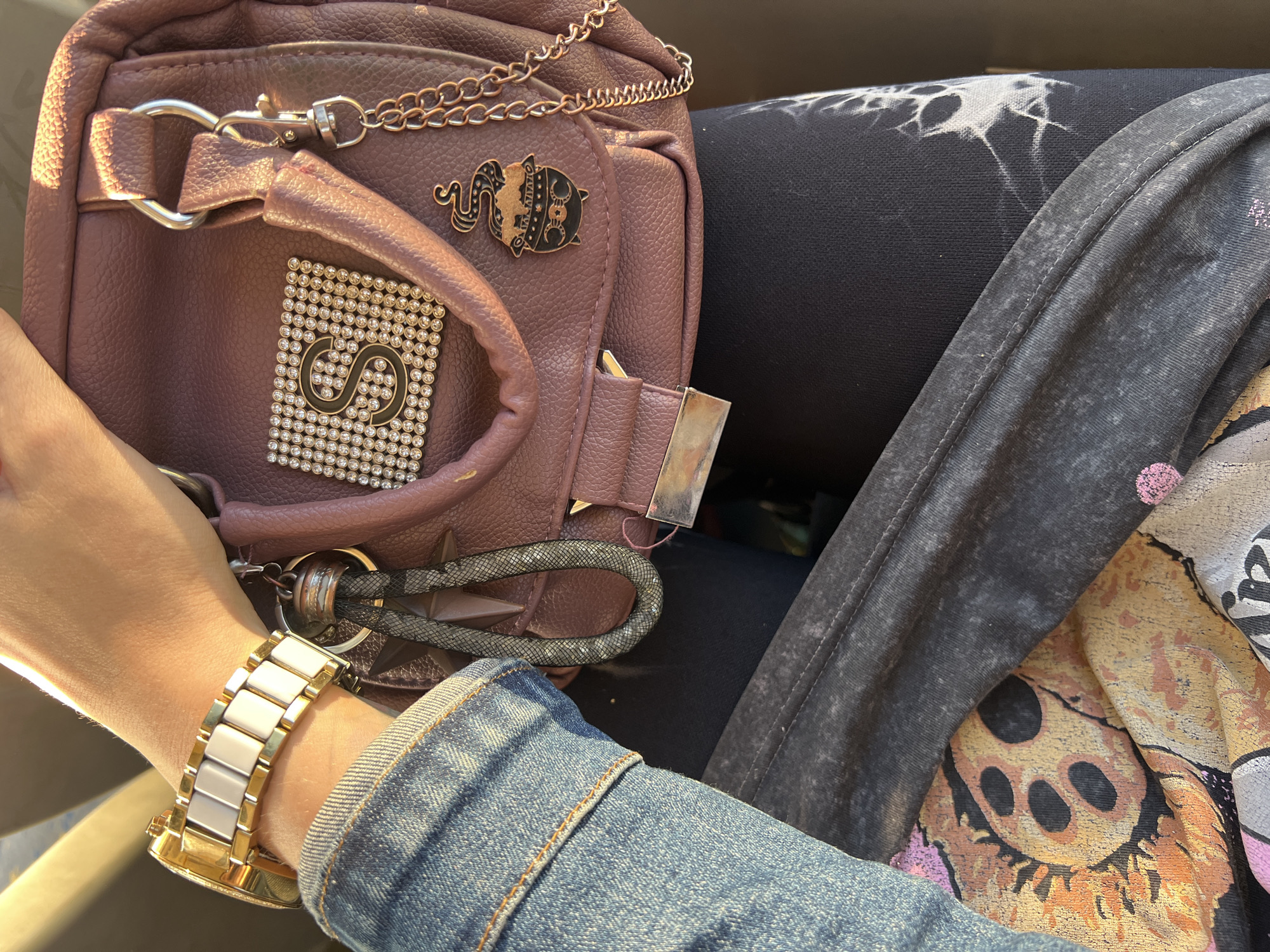 A close-up shot focuses on a person's arm and a small, dusty rose-colored handbag. The arm, adorned with a gold and white ceramic watch, is partially covered by a denim jacket. The handbag features a decorative patch with the letter "S" made of rhinestones, and a small enamel pin of a cauldron with smoke. A keychain with a braided grey strap hangs from the bag. The person is wearing dark leggings with a tie-dye pattern and a t-shirt with a graphic print of a cartoon skull. The background is out of focus, showing hints of yellow and brown, suggesting an outdoor setting with sunlight. The image is taken from a high angle, looking down at the subjects. The location is not specified in the image.