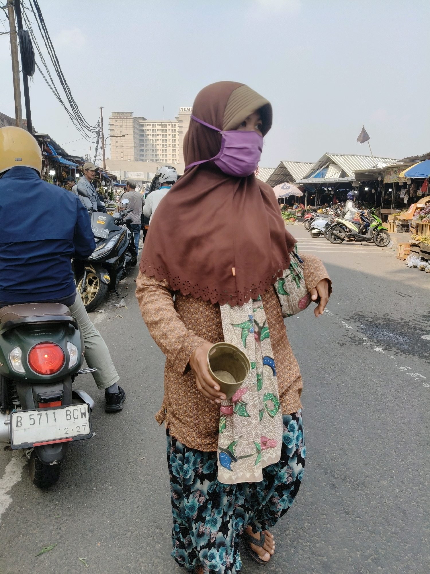 In the city of South Tangerang, Indonesia, a woman wearing a brown hijab and a purple face mask walks on a paved road, holding a brass cup in her right hand. She is dressed in a brown patterned shirt and a floral patterned skirt, with a fabric featuring a colorful bird motif draped over her left shoulder. Behind her, a person on a green scooter wearing a yellow helmet is visible. In the background, a bustling market with stalls and more people on motorcycles can be seen, with a large building labeled "NEWMARKET" in the distance. The sky is overcast.