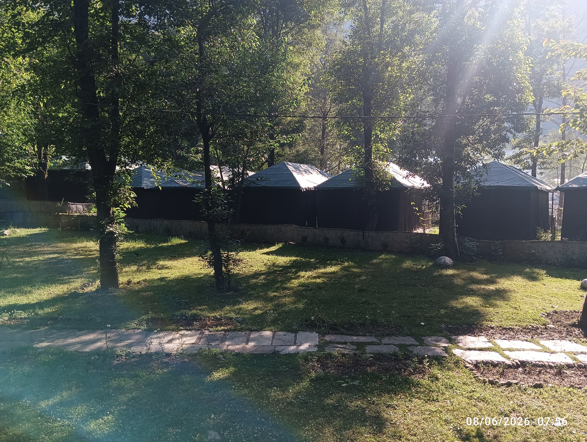 The image depicts a glamping or camping site with several dome-shaped tents set up in a grassy area surrounded by trees. The tents have dark fabric walls and light-colored conical roofs. A paved pathway made of square stones winds through the grass. Sunlight streams through the leaves of the trees, casting dappled shadows on the ground. The timestamp "08/06/2026 07:56" is visible in the lower right corner of the image. The scene appears to be a tranquil, outdoor setting. The location is identified as Goshal, India.