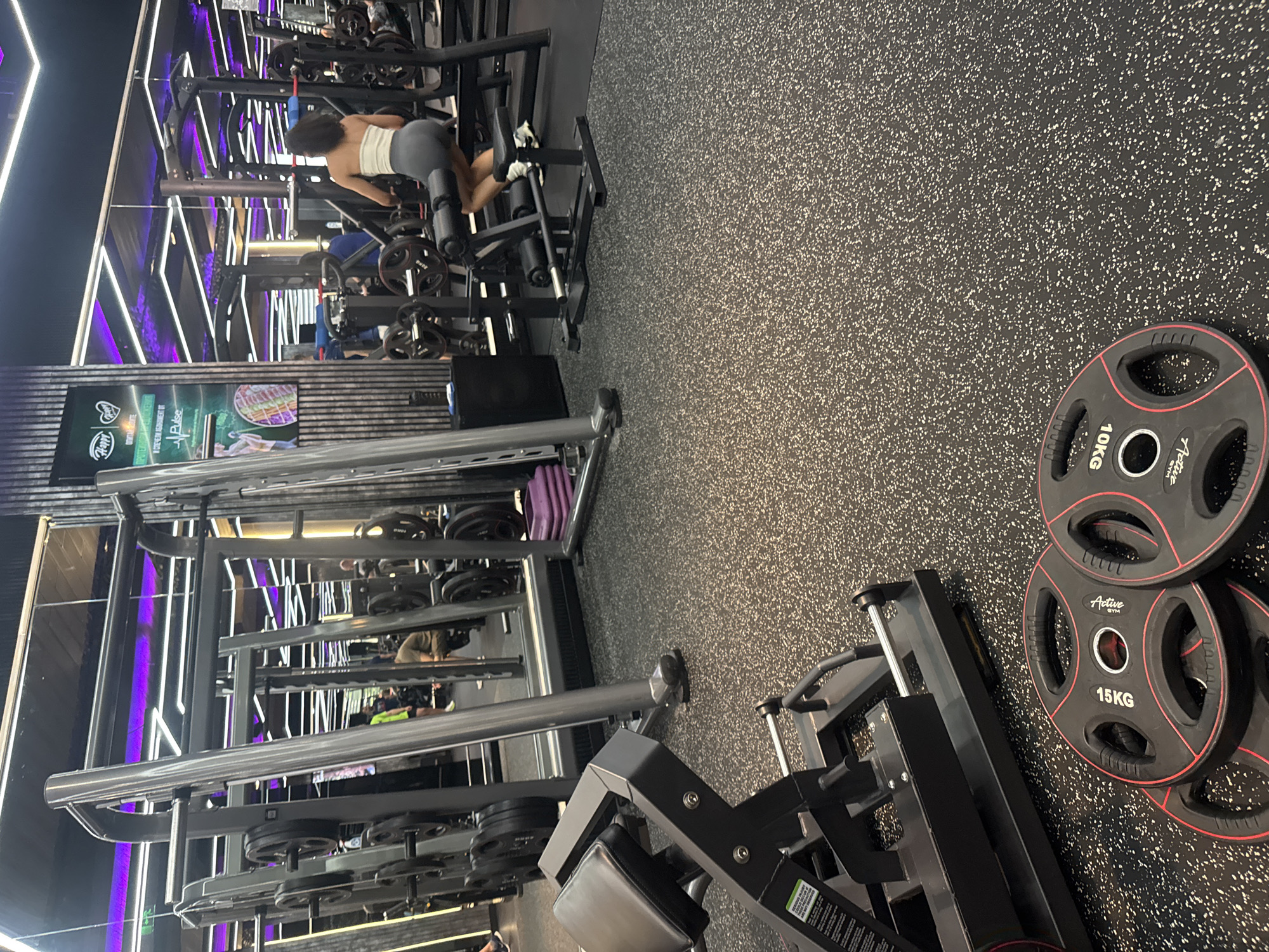 The image depicts the interior of a gym, likely in Plovdiv, Bulgaria, although no identifying details are visible. The gym features mirrored walls and numerous weightlifting machines. A woman in a white top and grey bottoms is using a hamstring curl machine in the background. In the foreground, a leg press machine is visible with weight plates, one marked "15KG" and another "10KG," lying on the floor nearby. The floor is black with white flecks. The gym has neon lighting. Other individuals and exercise machines are seen in the reflections and further back in the scene.