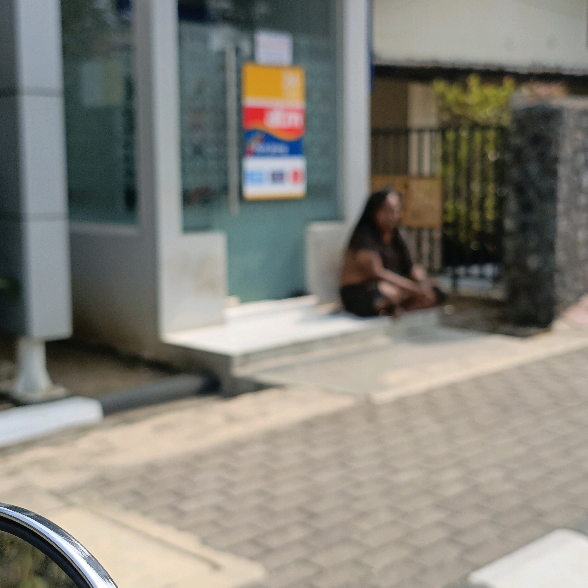 The image depicts an outdoor scene in Salatiga, Indonesia, with a blurred focus. In the background, a person is seated on a step in front of a building, which appears to be an ATM or a small commercial establishment, indicated by a signage with colorful panels, including yellow, red, and blue. The building has large glass panels. The ground in the foreground is paved with interlocking blocks. To the left, a reflective surface, possibly a motorcycle's rearview mirror, is visible at the edge of the frame. The overall impression is of a bright, sunny day.