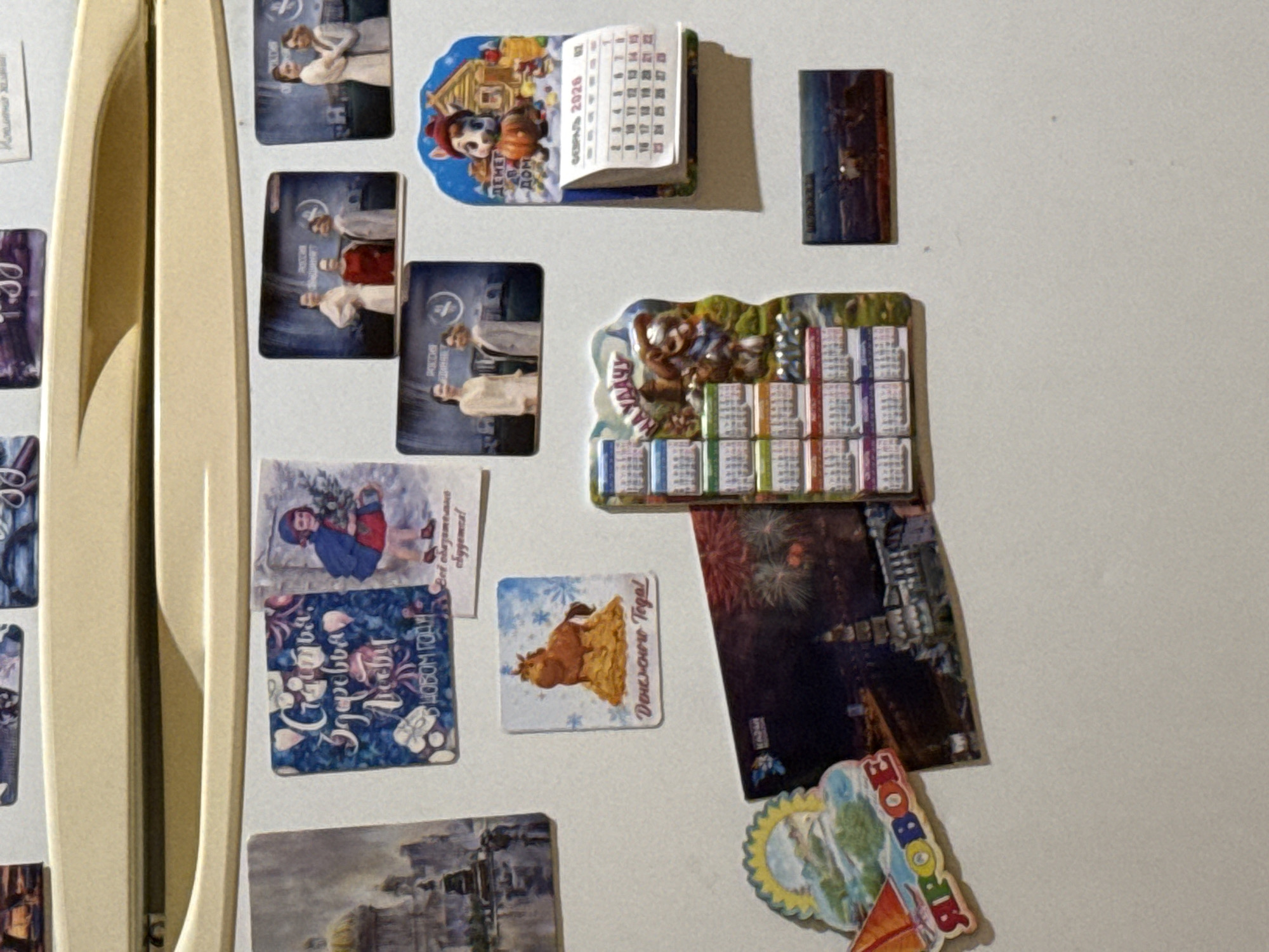 The image is a close-up, eye-level view of a refrigerator door covered in various magnets. The magnets are colorful and varied in their subjects, including a landscape painting, a festive holiday-themed magnet with Russian text, a magnet depicting a girl with a Christmas tree, a magnet of a horse on a pile of hay with Russian text, several rectangular magnets featuring people in formal attire, a magnet with a cartoon dog and a calendar, a magnet featuring a sailboat with Russian text, a magnet with fireworks over a cityscape, and a small rectangular magnet with an image of a landscape. One magnet is a calendar for March 2020. The refrigerator door is a light beige color and shows some signs of wear. There are no people visible in the image. The environment appears to be indoors, likely a kitchen. The city of Kholmsk, Russia is not depicted or referenced in this image.