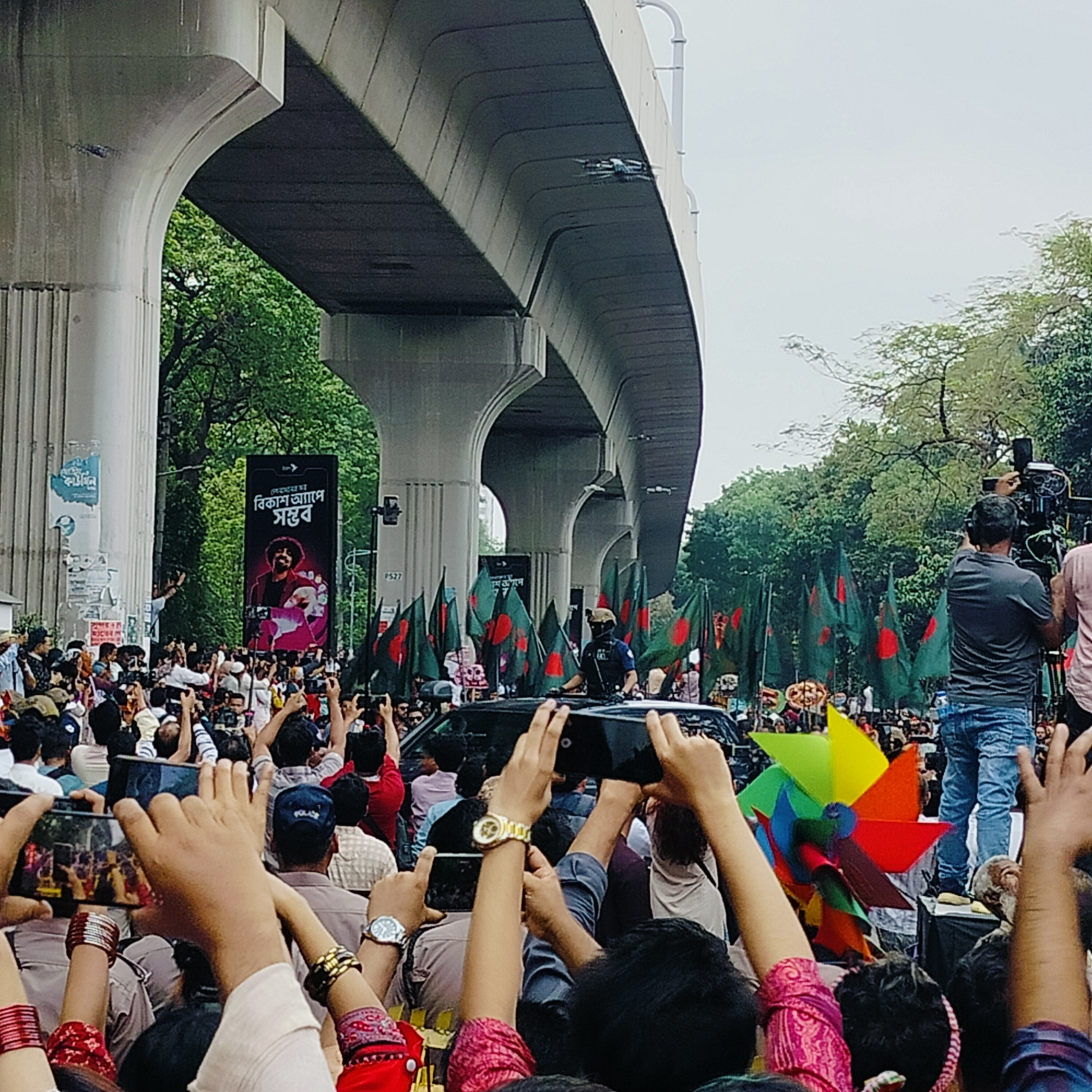 A large crowd of people is gathered in what appears to be Dhaka, Bangladesh, under an overpass. Many individuals are holding up their phones, seemingly taking photos or videos. Numerous green and red flags, indicative of the Bangladeshi flag, are visible throughout the crowd. At least one individual is on top of a dark car. A sign in Bengali is visible, and a colorful pinwheel can be seen in the foreground. A news cameraman stands on a platform capturing video of the event. The environment includes trees and overcast skies.