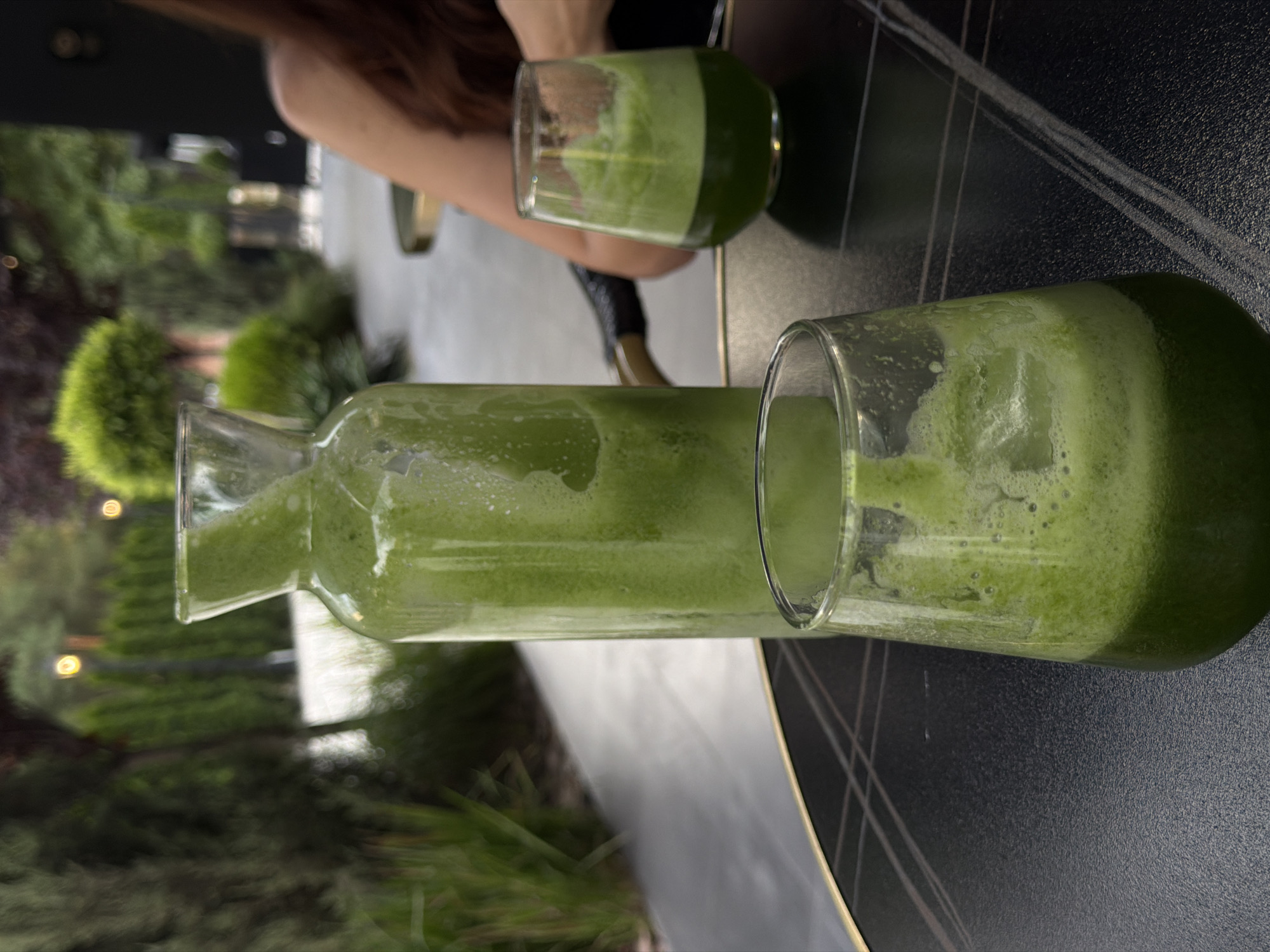 An outdoor dining setting in Bishkek, Kyrgyzstan, features a glass carafe and two glasses filled with a frothy green beverage on a dark patterned table. A portion of a woman's shoulder and upper arm is visible in the background, along with blurred greenery and outdoor lighting. The lighting suggests it is late afternoon or early evening. The overall scene is likely a cafe or restaurant patio.