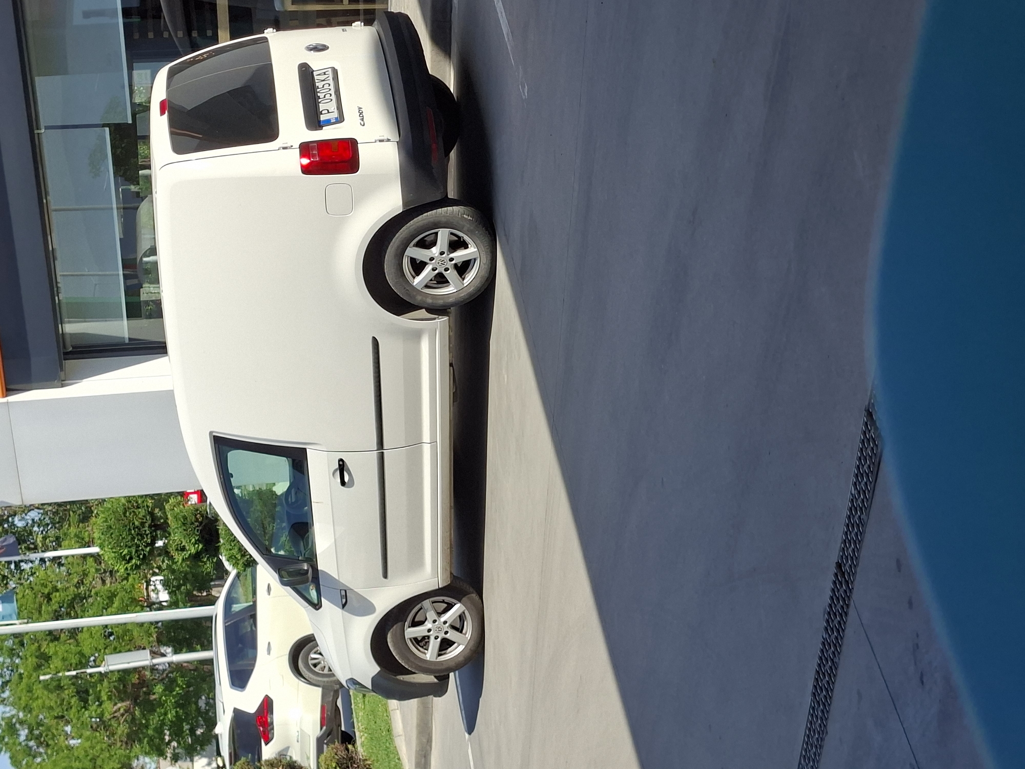 The image shows a white Volkswagen Caddy van parked on a gray concrete surface. The license plate is visible and reads "P 0505 KA." A building with a glass facade is visible in the background, along with some green trees and another parked car. Given the "P" on the license plate, it is probable that the image was taken in the city of Plovdiv, Bulgaria.