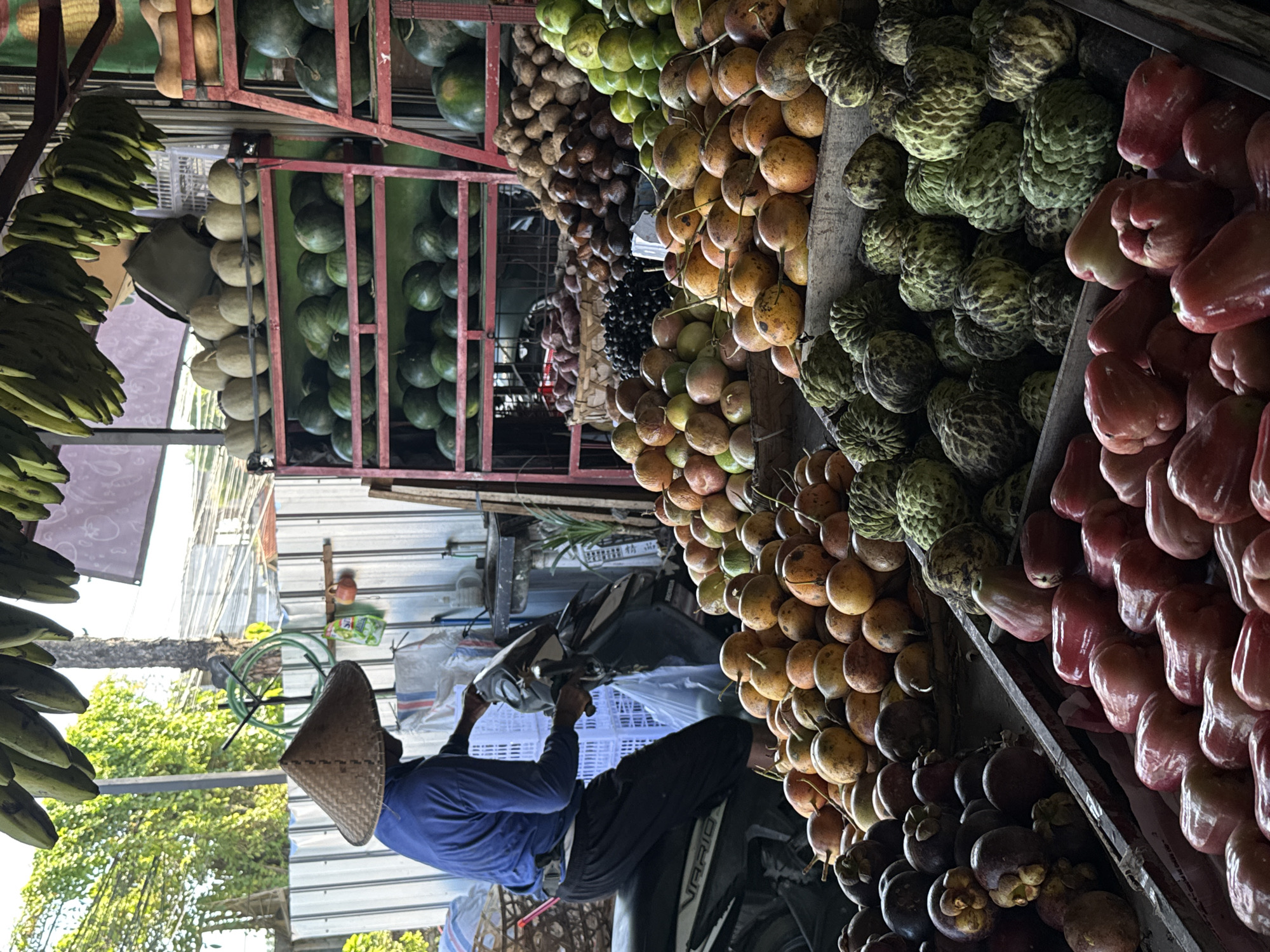 The image depicts a street-side fruit stall in North Kuta, Indonesia. A vendor wearing a conical Asian hat, a blue long-sleeved shirt, and dark pants stands beside a motorcycle. The stall displays a variety of fruits, including watermelons, bananas, passion fruit, custard apples, rose apples, and mangosteen. The fruits are stacked and arranged for sale within the stall's structure. The scene is set outdoors, with trees visible in the background.