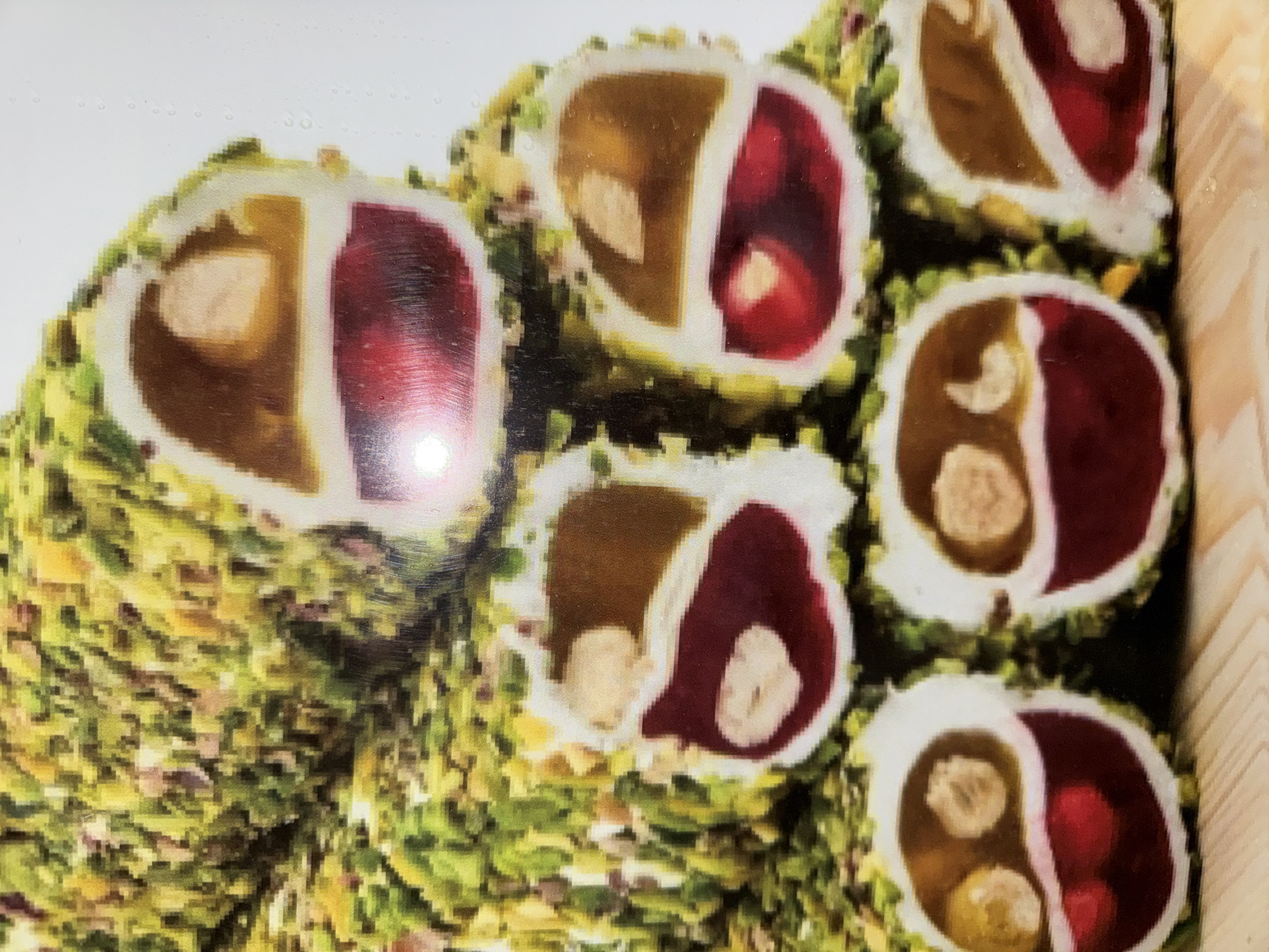 The image shows a close-up of Turkish Delight, specifically the pistachio-covered variety. Each piece is cut in half, revealing a two-tone interior, divided into a reddish section and a brownish section. The reddish section contains red fruit pieces, while the brownish section contains nuts. Based on the common presence of Turkish Delight in tourist areas, and the prevalence of this confection in Alanya, Turkey, it can be surmised that this photograph could plausibly have been taken in that city.