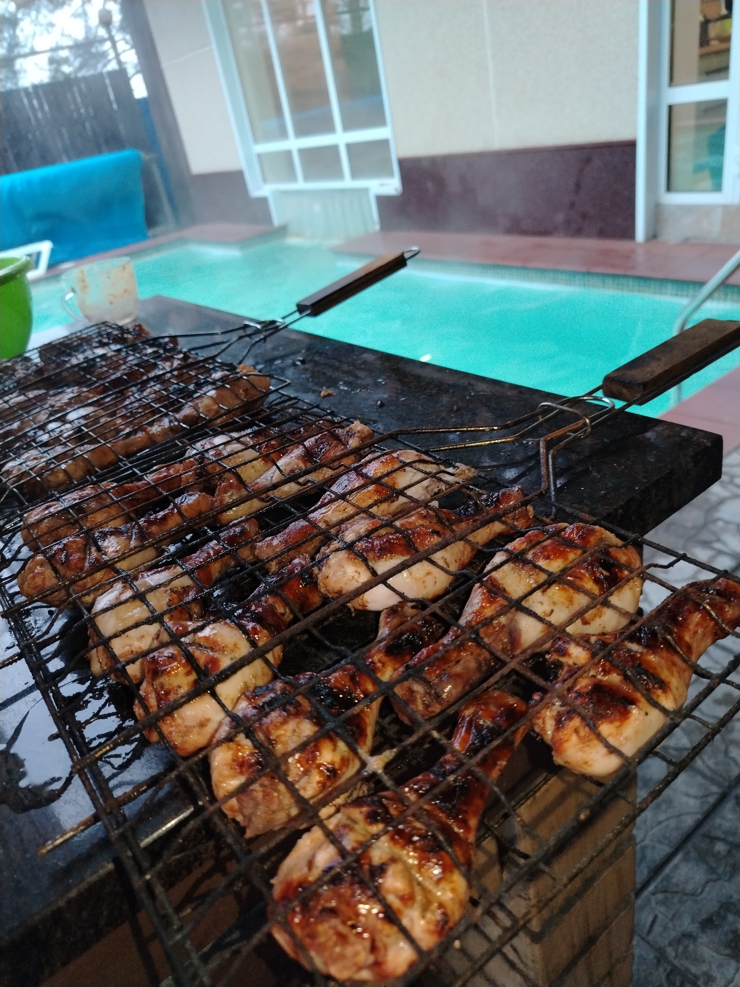 The image captures an outdoor scene in Chaikovsky, Russia, centered on chicken being grilled by a swimming pool. The main subjects are several pieces of chicken, primarily drumsticks and other cuts, arranged on two metal grilling baskets. The chicken is well-cooked, showing a golden-brown hue, some distinct char marks from the grill, and a glistening surface, suggesting a marinade or sauce. The grilling baskets feature wooden handles and are placed over a dark, solid grill or brazier.
To the left of the grill, a partial view of a green bucket or bowl and a clear glass mug can be seen. The ground around the grilling area is paved with light-colored tiles. In the background, a striking light blue swimming pool with clear water is prominent, with some steam possibly rising from its surface. A blue towel or fabric is visible near the pool's edge. Beyond the pool, modern buildings with large windows are discernible, indicating a residential or resort setting. The lighting is diffused, suggesting an overcast day, late afternoon, or early evening. The scene evokes a sense of leisurely outdoor dining. There are no people visible, and no visible text is present.