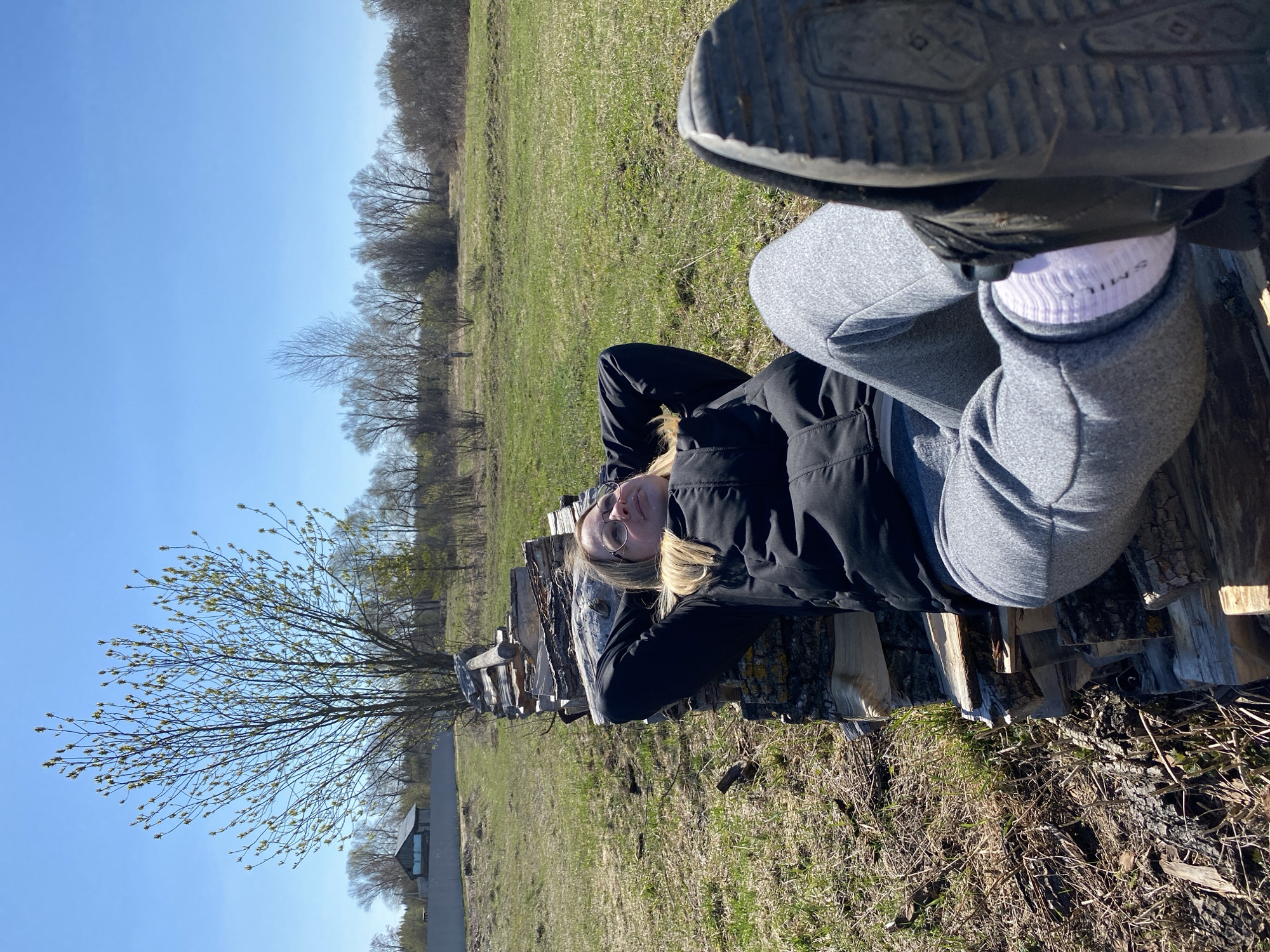A woman is lying on a stack of logs in an outdoor setting. She is wearing a black jacket, grey sweatpants, and white socks with the word "SMILE" printed on them. She has blonde hair and is wearing glasses. Her shoes are propped up in the air.
The setting appears to be a field or grassy area with trees in the background. The sky is clear and blue. There is a building visible in the distance.
Without any information regarding the specific environment in Kauchuk Stantsiya, Russia, the setting cannot be specified further.