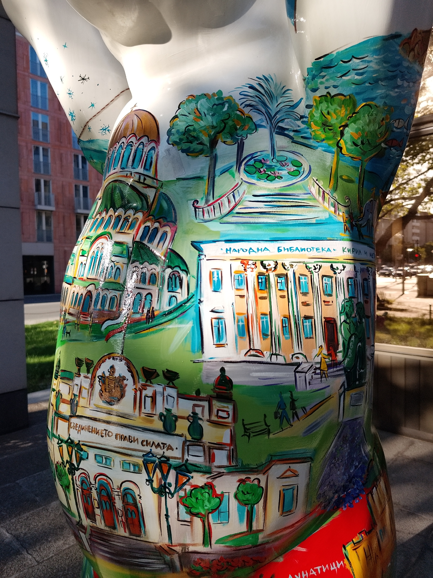 A brightly painted sculpture stands outdoors, depicting several architectural landmarks. The top half features a multi-domed building, likely a cathedral, alongside a scene with trees, a pond with lily pads, and a body of water in the background. Further down is a building with prominent columns, and the text "НАРОДНА БИБЛИОТЕКА КИРИЛ И МЕТОДИЙ," which translates to "National Library Cyril and Methodius." Below that is another building with a coat of arms and the text "СЪЕДИНЕНИЕТО ПРАВИ СИЛАТА," which translates to "Unity Makes Strength." In the foreground, there are street lights and trees, as well as the text "АУНАТИЦИ." The surrounding environment includes a modern brick building in the background and a blurred view of a street with a reflection of trees and buildings on a glass surface to the side. The sculpture appears to be in a public urban setting. The location is inferred to be Sofia, Bulgaria, based on the landmarks depicted.