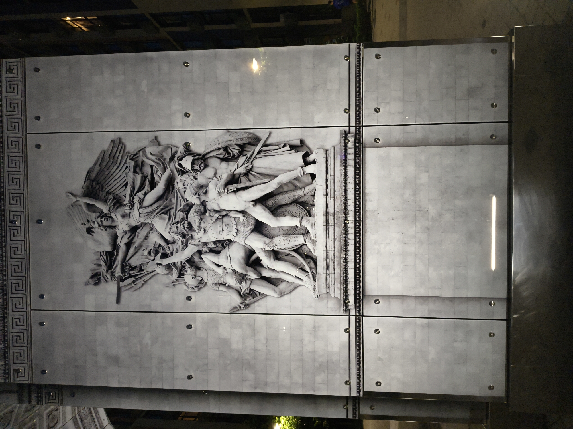 The image captures a nighttime view of a large, monochrome bas-relief sculpture mounted on a stone-tiled facade. The sculpture depicts a dynamic scene of figures engaged in combat, with a winged female figure at the top and several muscular male figures below, some armed with swords and shields. The facade is divided into two vertical panels and features a Greek key pattern border at the top. The overall style is neoclassical. The setting appears to be an urban environment, with a building visible to the right and faint street lighting. The sculpture is likely a decorative element on a public building or monument. The text provided states the location is in Kyiv, Ukraine.