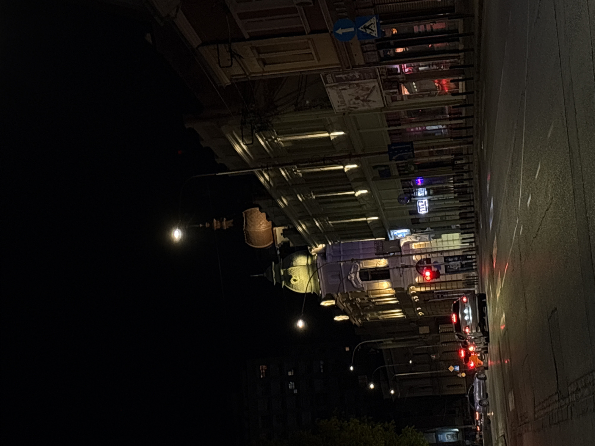 The image shows a nighttime street scene in Kragujevac, Serbia. The camera is positioned looking upwards along a street lined with buildings. Streetlights illuminate the architecture, including a dome-shaped structure, while traffic lights are visible at an intersection further down the street. Cars with illuminated taillights are stopped at the intersection. The left side of the image is predominantly dark, suggesting it is an area without direct illumination. Road markings are visible on the street surface. Street signs are also visible.
