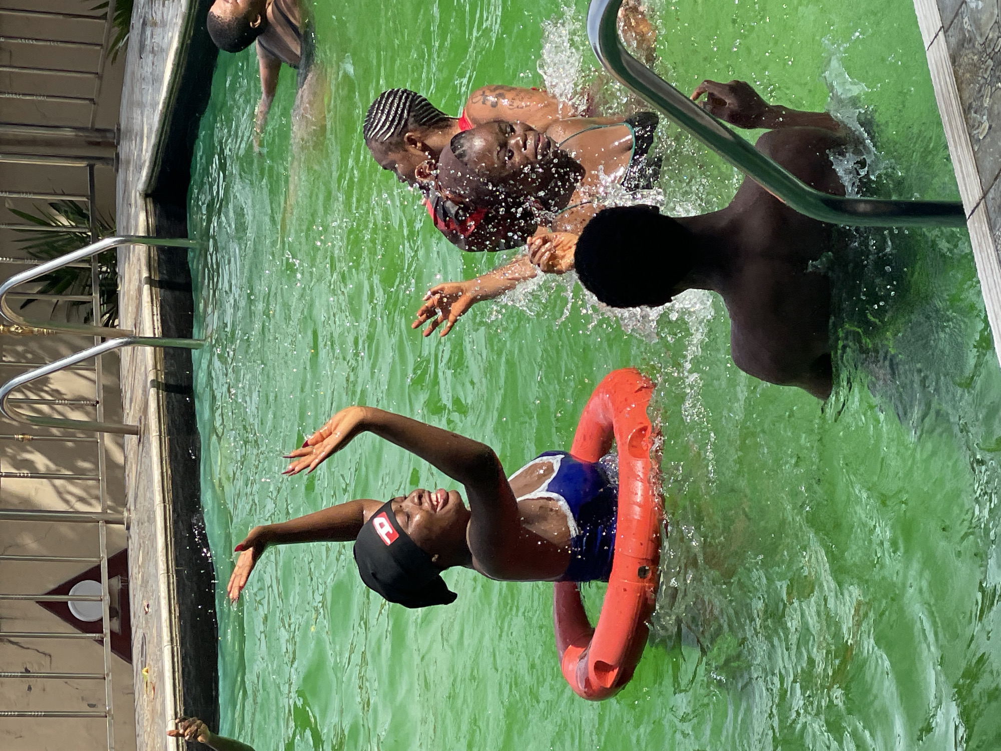 A group of people are enjoying a sunny day in a bright green swimming pool. A woman in the foreground wears a blue and white swimsuit and a black headscarf, smiling with her arms raised. She is partially encircled by a red float. To her right, two other women are interacting in the water, one with braided hair. Further in the background, another person is visible swimming. The pool area is surrounded by a tiled deck and a metal fence. Sunlight reflects off the rippling water, creating a vibrant atmosphere. The scene appears to be a leisure activity, possibly a party or gathering, at a residential or public pool.