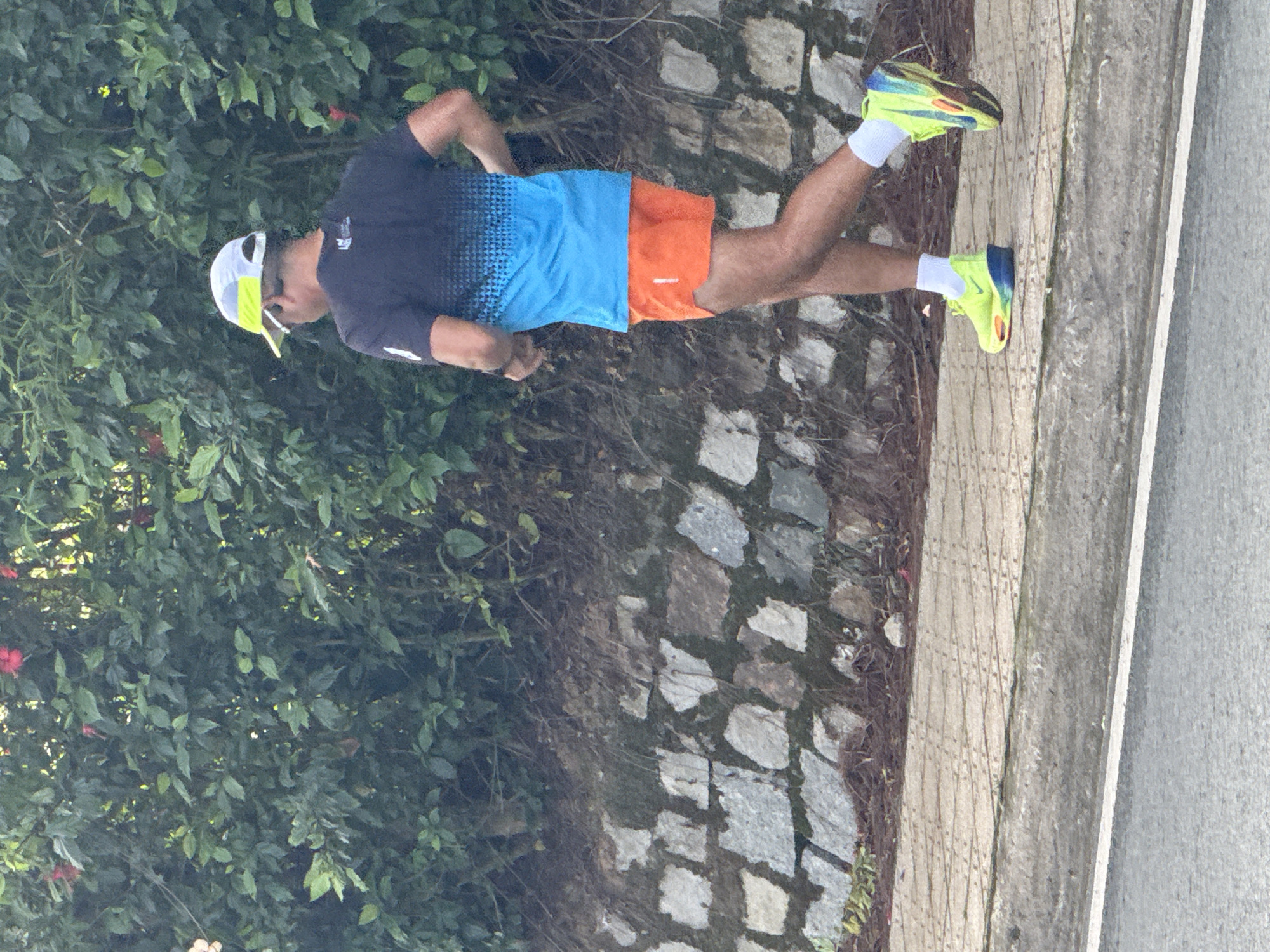 A man is running on a paved sidewalk. He is wearing a dark blue and bright blue t-shirt, bright orange shorts, white socks, and neon green and blue running shoes. He also wears a white and yellow baseball cap. The man is running alongside a wall made of various sized stones. Behind the wall is a lush green bush with some red flowers. The sidewalk is adjacent to a road. The image appears to have been taken in Da Lat, Vietnam, based on the presence of similar stone walls and foliage seen in the city.