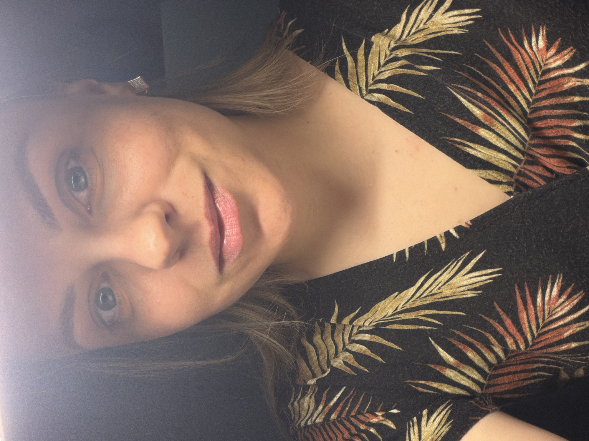 A close-up of a woman's face and upper torso fills the frame. The woman has light skin, light brown hair, and light eyes. She is wearing a dark-colored top with a pattern of gold and red-brown palm leaves. A bright light source is to the left of the frame, casting a glare. The background is dark and indistinct. The photo does not provide enough information to determine the location in Votkinsk, Russia.