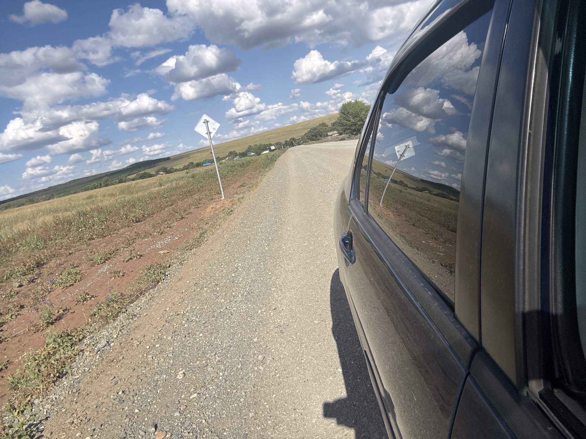 A vehicle travels on a gravel road in Васильевка, Russia. The car, a dark-colored sedan, is seen from the side, with the driver's side door and window in the foreground. The window reflects the blue sky and white clouds, as well as a diamond-shaped sign on a pole. The road is lined with dry grass and reddish-brown soil on the left side, with a gentle slope of green hills in the background. Small houses and trees are visible on the hills in the distance. The sky is clear with scattered cumulus clouds.