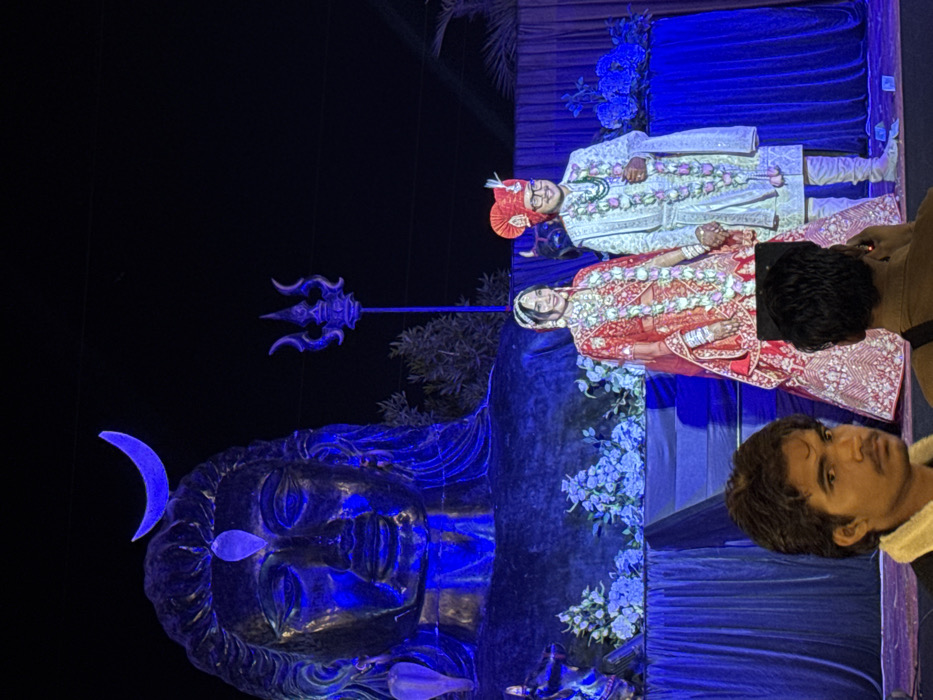 A color photograph shows a couple dressed in traditional wedding attire, posing in front of a statue of Shiva. The bride wears a red saree, and the groom wears a light-colored suit and a red turban. Both are adorned with garlands. A trident is also visible. The statue of Shiva is illuminated with blue light. Two onlookers are present, one with a phone and the other looking into the camera.