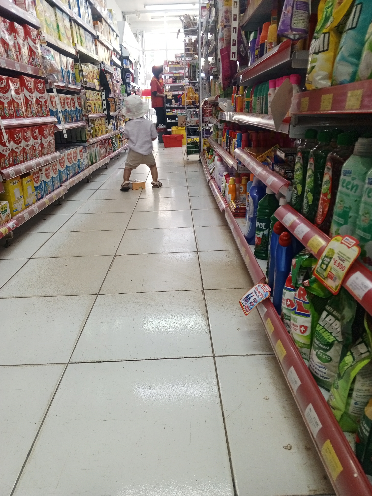 A young child wearing a white hat and shorts stands in an aisle of a brightly lit supermarket. The child is facing away from the camera, looking down an aisle lined with shelves stocked with various products. To the left, shelves are filled with boxes of what appears to be powdered milk. To the right, shelves hold bottles of cleaning supplies and detergents. In the background, a person in an orange shirt and dark pants stands near the end of the aisle. The floor is tiled in a light color, and the overall environment suggests a retail space. The location is identified as a supermarket in Talun Kenas, Indonesia.