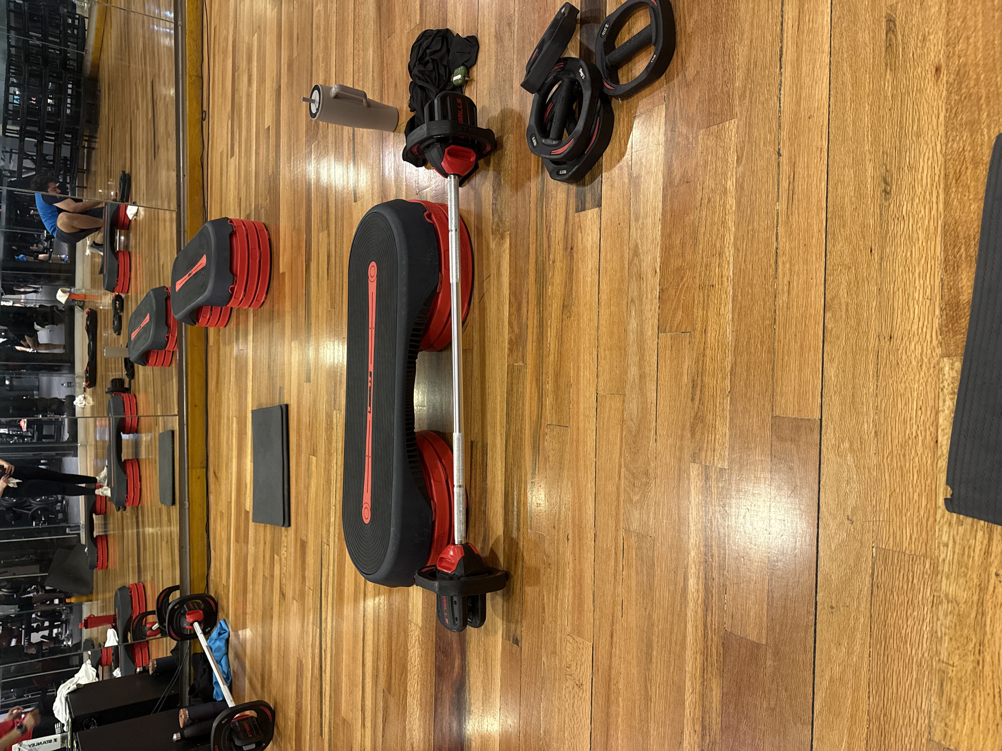 The image shows a gym floor with various exercise equipment laid out, including a barbell with weights, exercise steps, a foam roller, and weight plates. The floor is made of wood. In the background, a mirror reflects the gym setting, showing people exercising. The location is likely a gym in an urban setting, potentially in Puebla, Mexico, as specified.