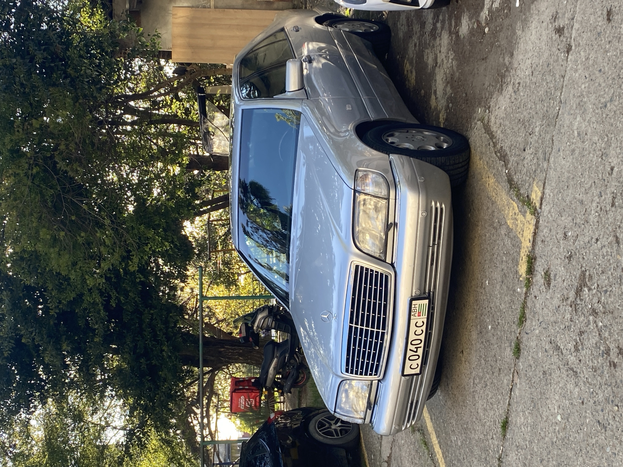 A silver Mercedes-Benz sedan, likely a W140 S-Class model, is parked outdoors on a concrete surface during bright daylight. The car is angled towards the viewer, showing its front grille, headlights, and the left side. Sunlight filters through dense, leafy green trees that dominate the background and left side of the scene.
On the front of the Mercedes, a license plate is clearly visible, displaying the characters "C040 CC" along with the flag of Abkhazia and the country code "ABH." To the left of the Mercedes, a black scooter or moped is parked, and parts of a black car are visible. The front bumper of a white car can be seen in the upper right background. The concrete pavement beneath the vehicles features faded yellow painted lines and some patches of grass and moss. No people are visible in the image. The setting appears to be a parking area or street, consistent with the urban or suburban environment of Sochi, Russia, which borders Abkhazia.