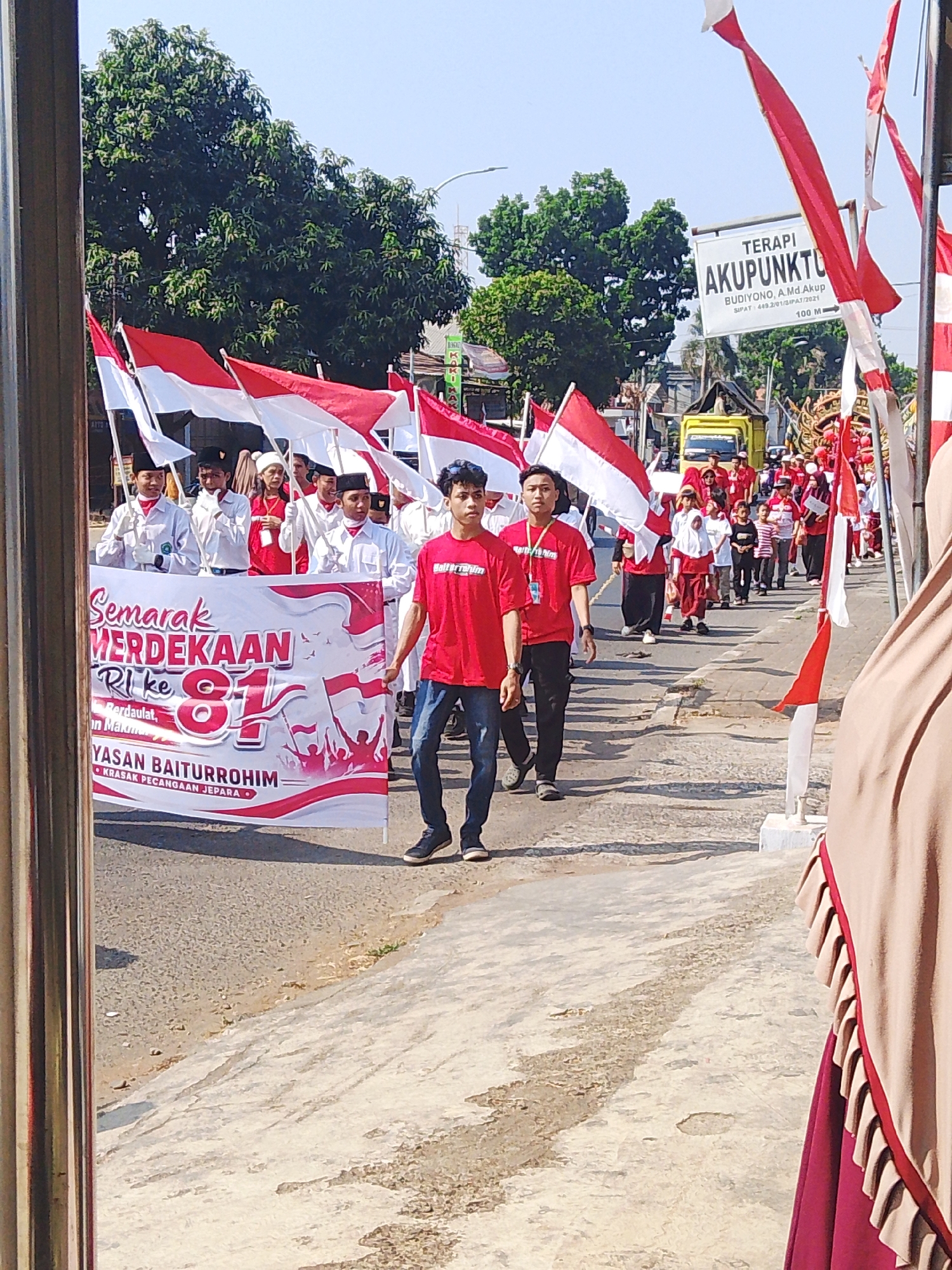 A group of people, some holding Indonesian flags, are participating in a parade on a sunny day. The parade appears to be celebrating Indonesia's Independence Day, indicated by the banner with "Semarak MERDEKAAN RI ke 81" (Grand Celebration of Indonesian Independence 81st) written on it. The people are dressed in red and white attire, consistent with the national colors. Some individuals are wearing red t-shirts with "Baiturrohimp" printed on them, and others are in white uniforms with black caps. A yellow truck is visible in the background, along with other participants and spectators. The scene takes place on a paved road with trees and buildings lining the sides. A signpost with the text "TERAPI AKUPUNKTU" and directional information is also visible. This event is likely taking place in Kalinyamatan, Jepara, Indonesia, as suggested by the text on the banner.