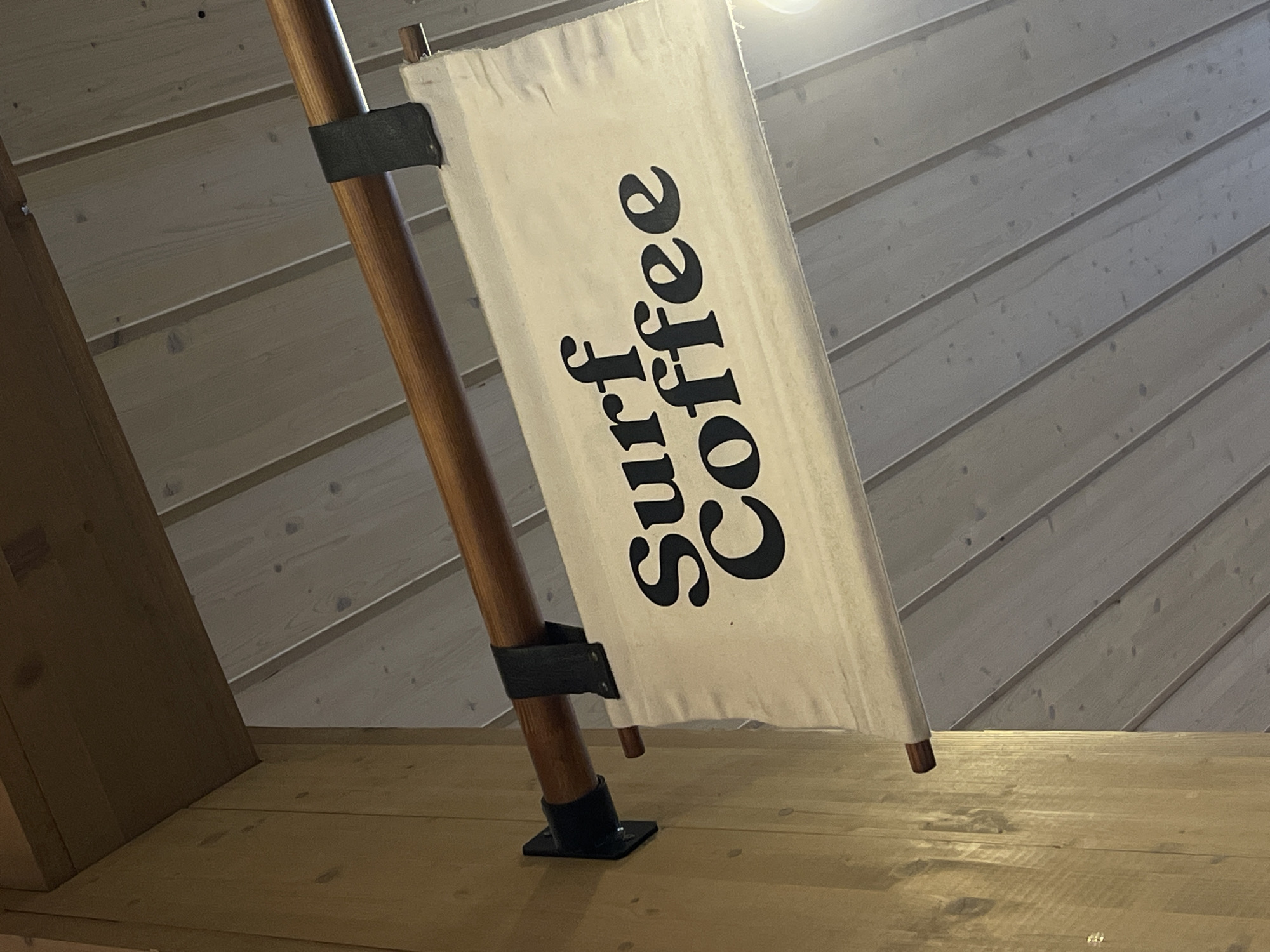 The image shows a sign that says "Surf Coffee" in black, stylized font on a cream-colored fabric banner. The banner is hung from a wooden pole suspended by black straps. The pole is attached to a wooden beam. The sign appears to be indoors, with a wooden paneled wall on the left and a wooden slatted ceiling above. The scene is well-lit, with a bright light source visible in the lower right corner. There are no people visible in the image. There is no information in the image that indicates the location is Mistolovo, Russia.
