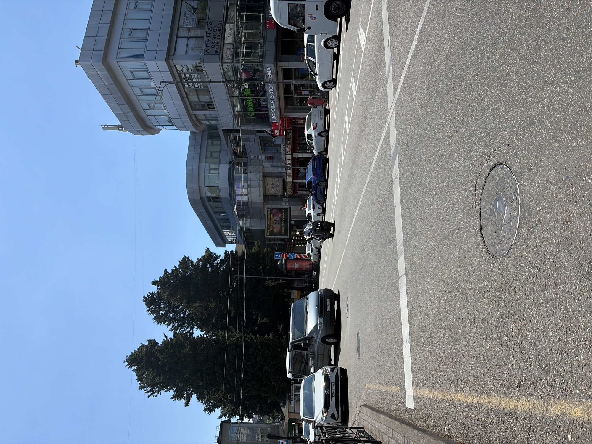 This is a medium-distance, eye-level, outdoor, daytime shot of a street intersection in Sochi, Russia. On the right side of the frame, a white passenger van, likely a Mercedes-Benz Sprinter, is parked. In the foreground, the asphalt street shows clear lane markings, including white lines and a yellow line along the curb. A circular manhole cover is visible in the center of the road. Several vehicles are on the street, including a white Volkswagen sedan with license plate T125AT193, a gray Ford Transit van with license plate C328VX59, a dark blue BMW sedan, and several other white cars. A motorcyclist in a helmet and white shirt is riding on the road. In the background, there are multi-story buildings with modern architecture, featuring glass facades. Trees are visible behind the buildings. The sky is clear and blue. The scene depicts a typical urban environment with moderate traffic.