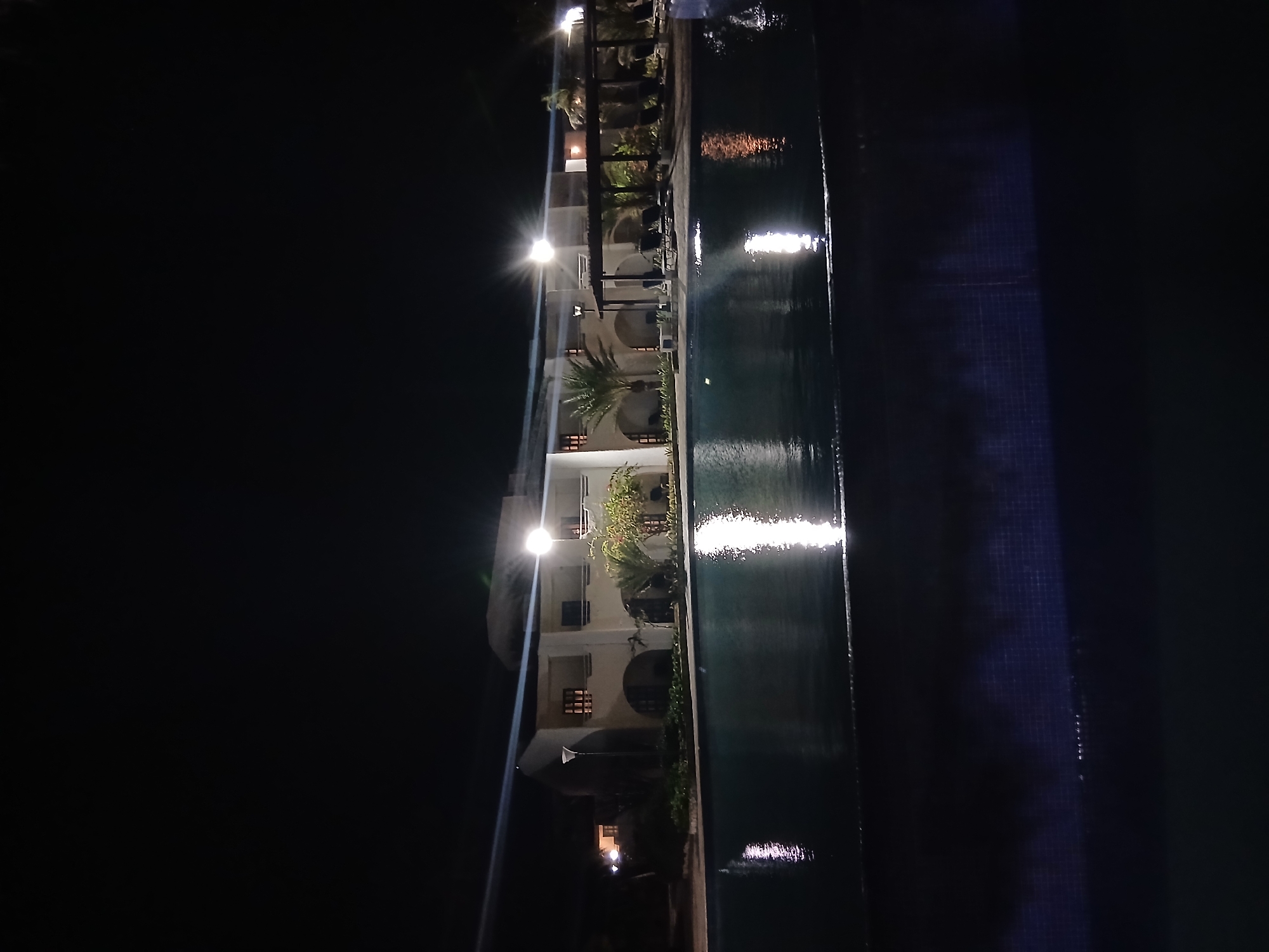 The image displays a nighttime scene of a resort property located in Altagracia, Venezuela. A two-story building with a light-colored facade, featuring multiple arched ground-floor openings and rectangular upper-level windows, stands across a large, dark-water swimming pool. The building's exterior is illuminated by several bright artificial lights, including a tall, modern lamppost on the left, creating distinct, elongated reflections on the pool's surface. Palm trees and other dense green foliage are distributed around the building, which also includes a covered outdoor area on the right, furnished with multiple dark lounge chairs. The upper portion of the image shows a completely dark night sky. No individuals are present in the scene.