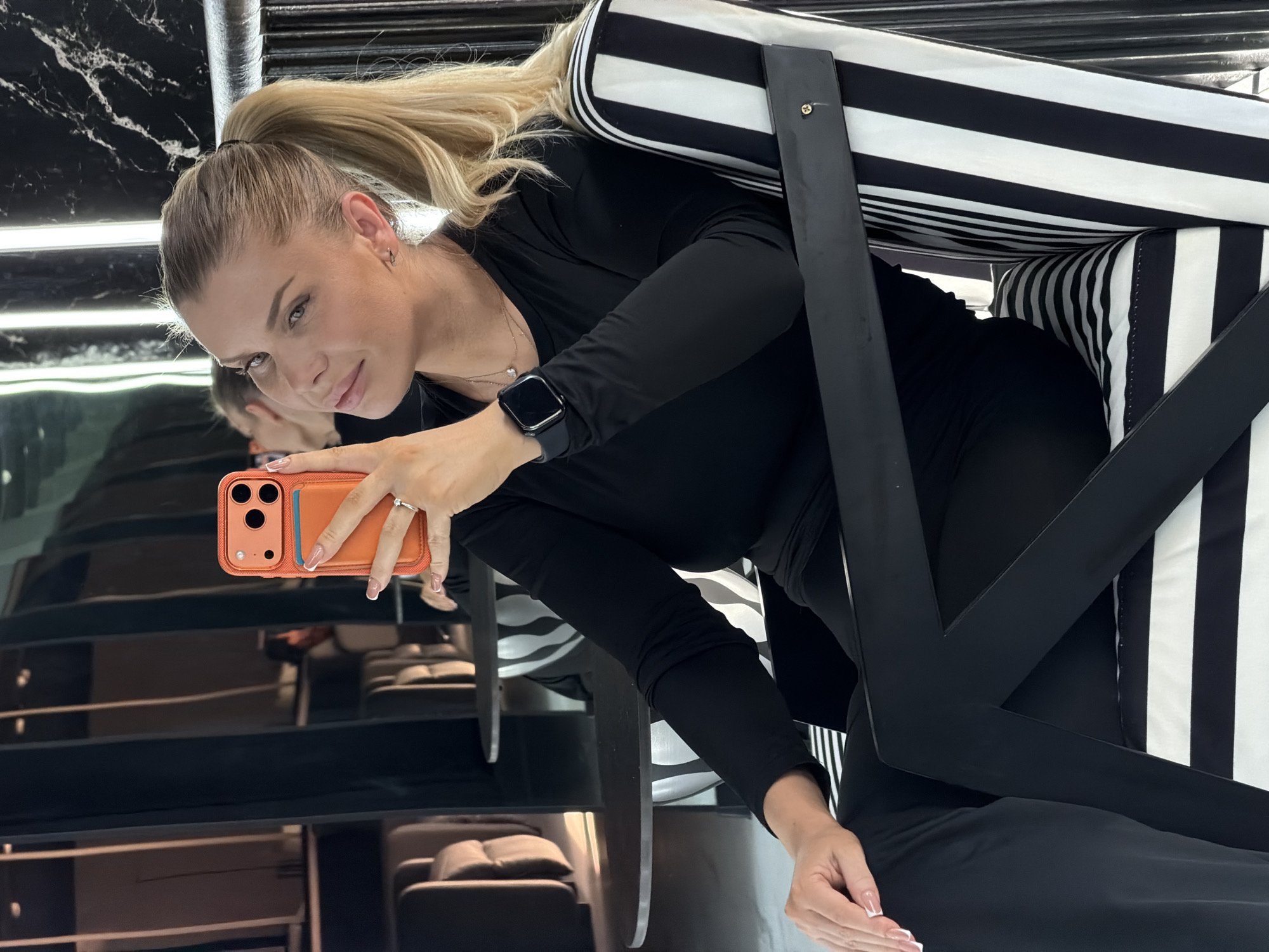 A woman with blonde hair in a ponytail is taking a selfie in a modern interior. She is wearing a black long-sleeved shirt and black pants, and is sitting in a chair with black and white stripes. She holds an orange phone in her right hand. The background appears to be a lounge area with reflective surfaces and architectural lighting. The location is not specified in the image.